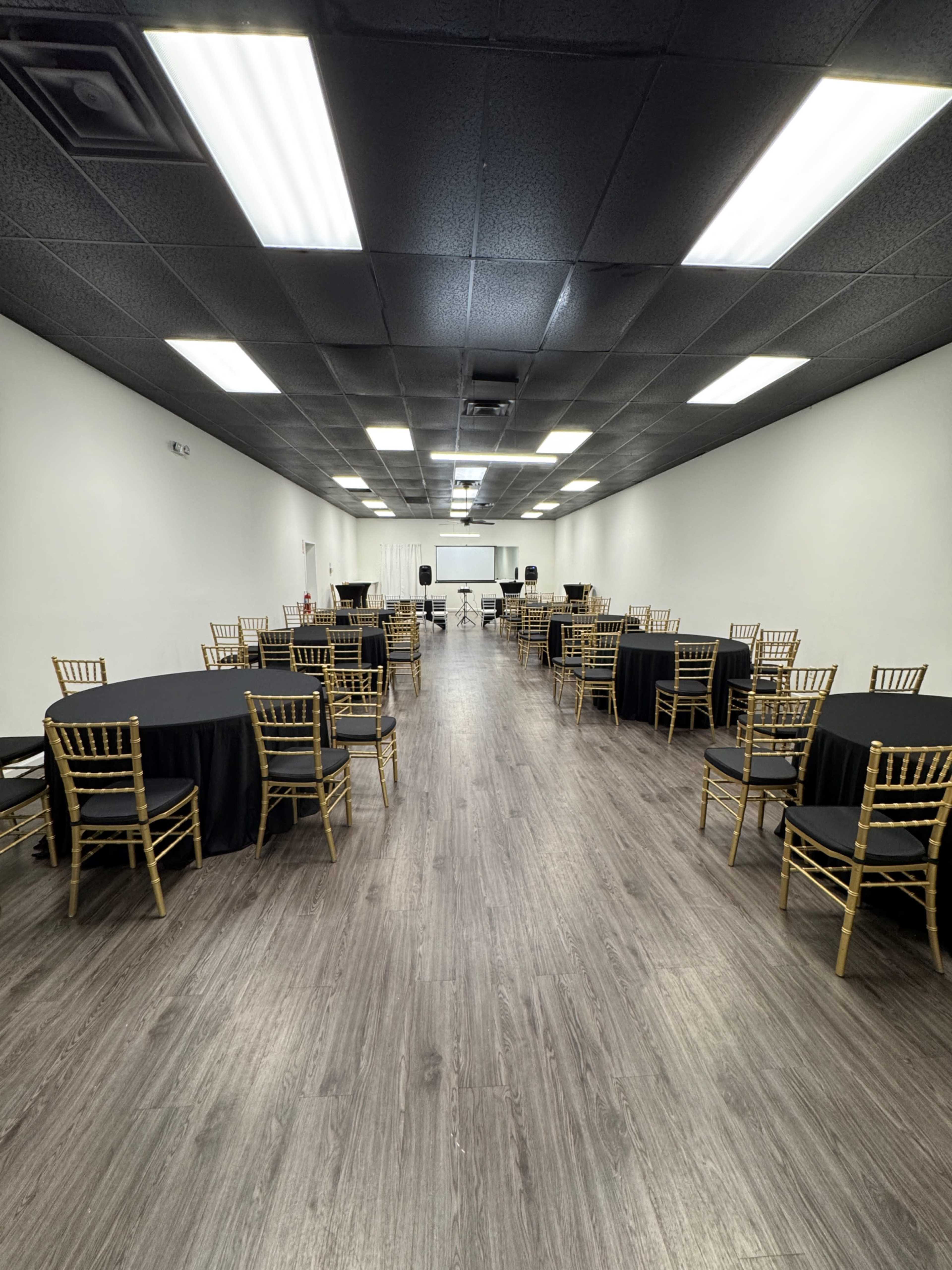 The image shows a large event space with several round tables covered in black tablecloths and gold chairs arranged in a straight line down the center.