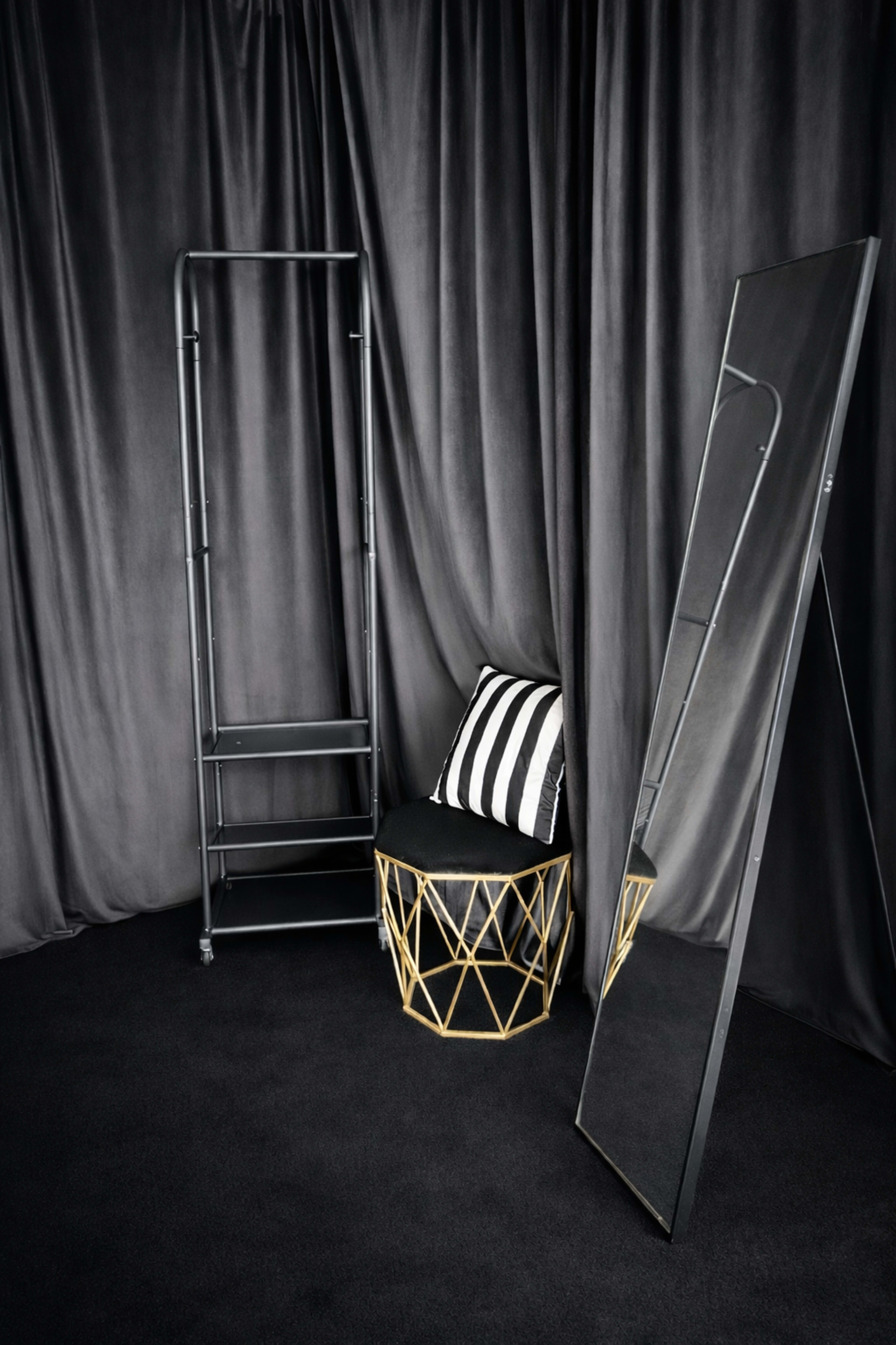 The image shows a modern setting with a reflection mirror placed next to a geometric gold and black stool, against a backdrop of dark curtains.