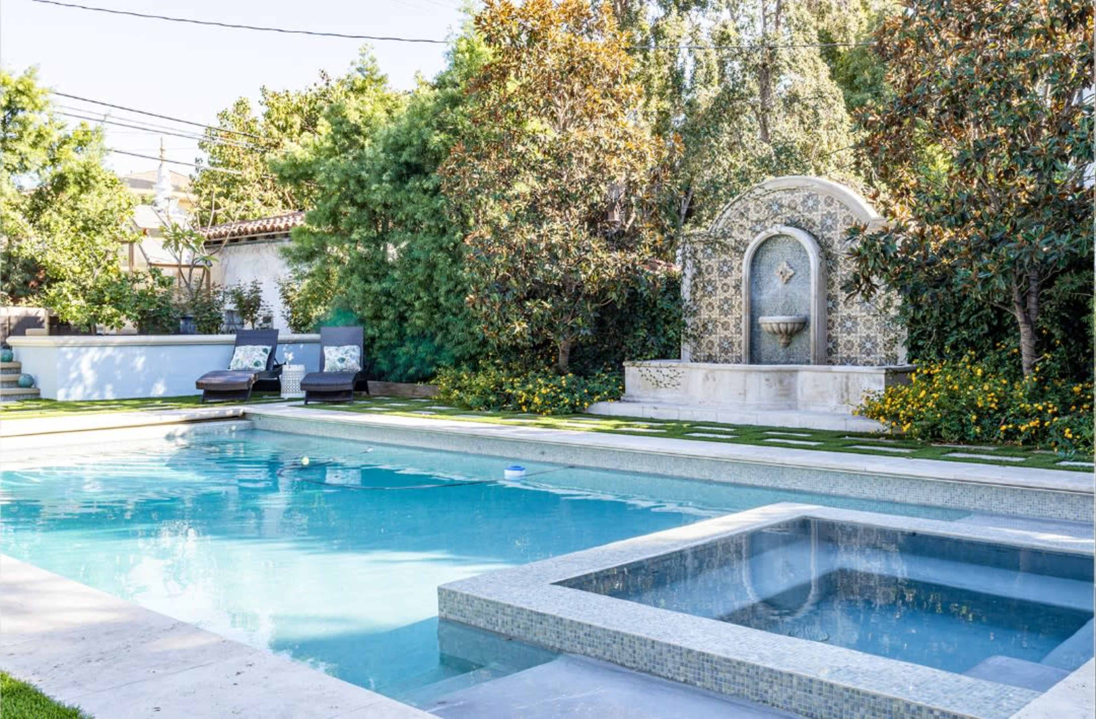 The image shows a backyard scene featuring a rectangular swimming pool with a spa, surrounded by greenery and a decorative fountain.