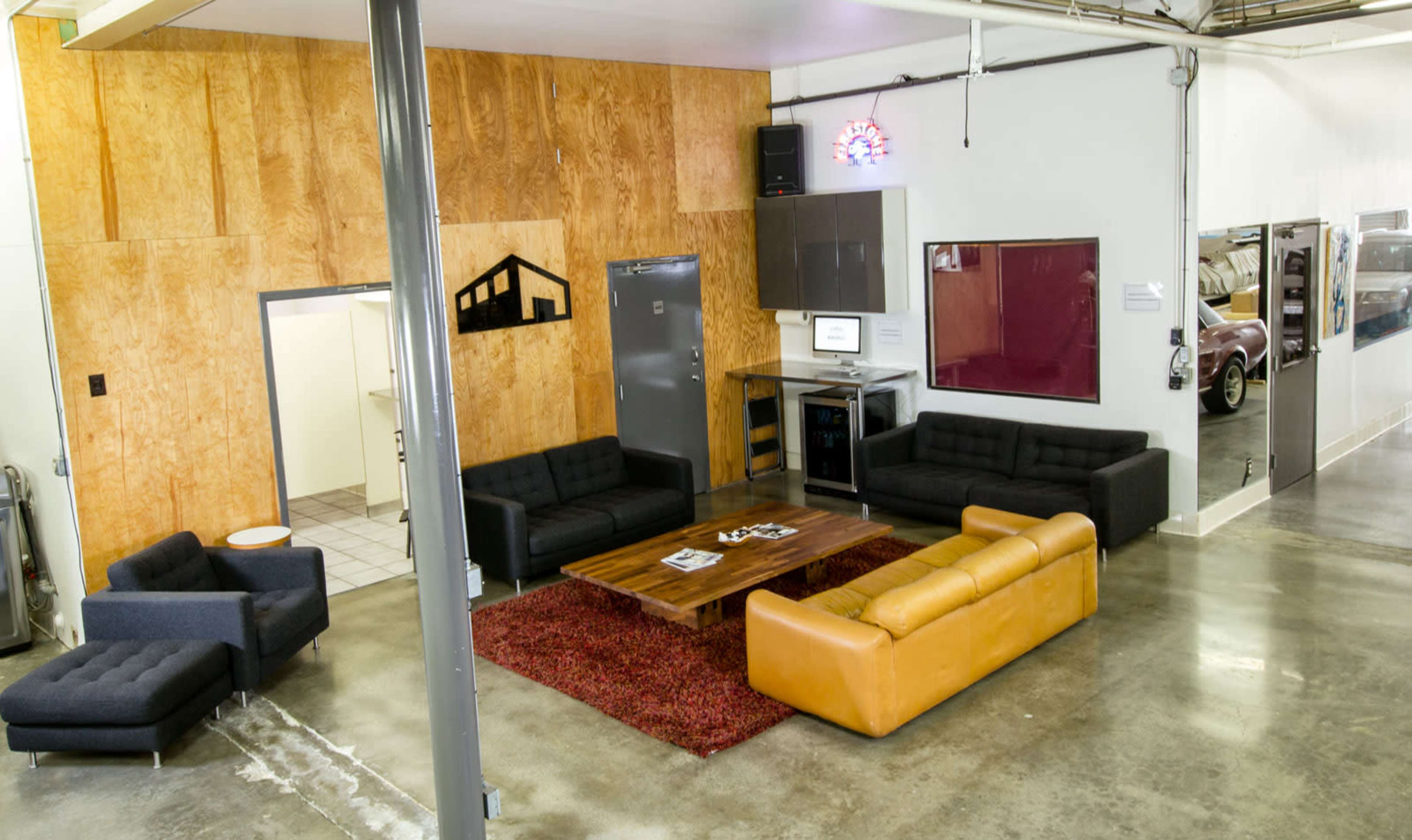 The image shows a modern lounge area with dark sofas and a light leather couch, a wooden coffee table, and a wall featuring wood paneling, with a kitchen visible in the background.