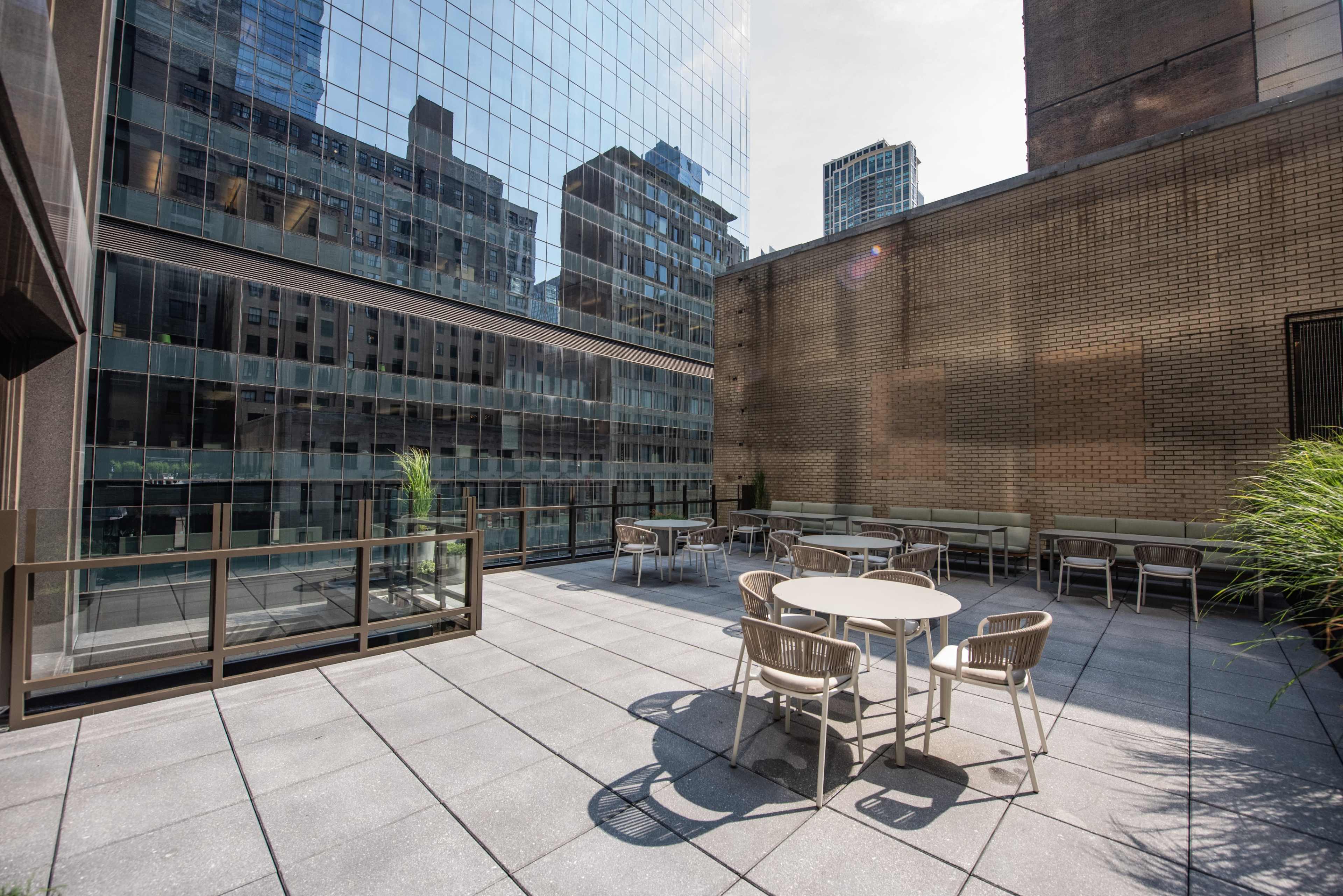 The image shows a rooftop terrace with tables and chairs surrounded by tall buildings in an urban setting.