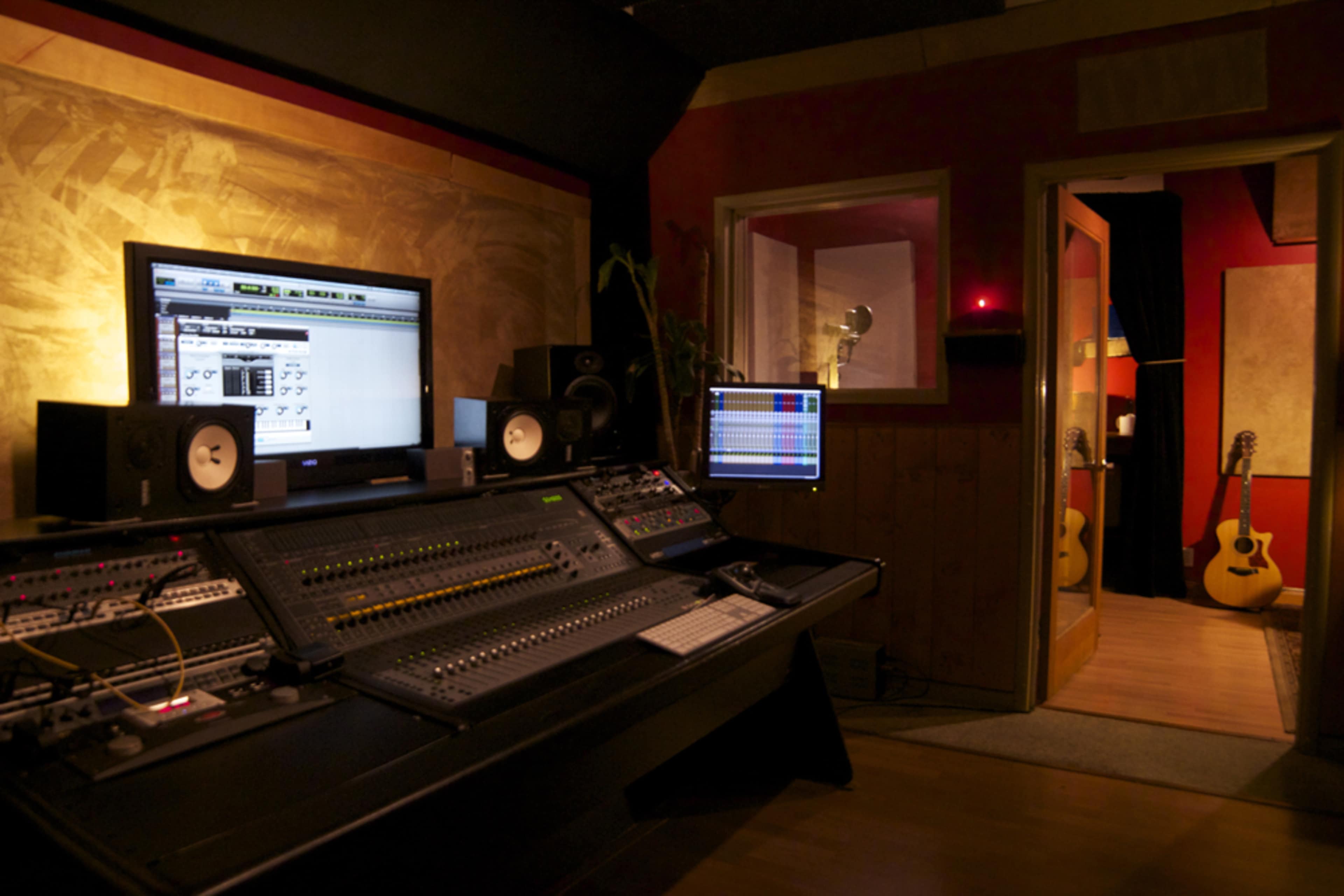The image shows a professional music recording studio with a control console, computer monitors, and a hallway leading to a room with guitars.