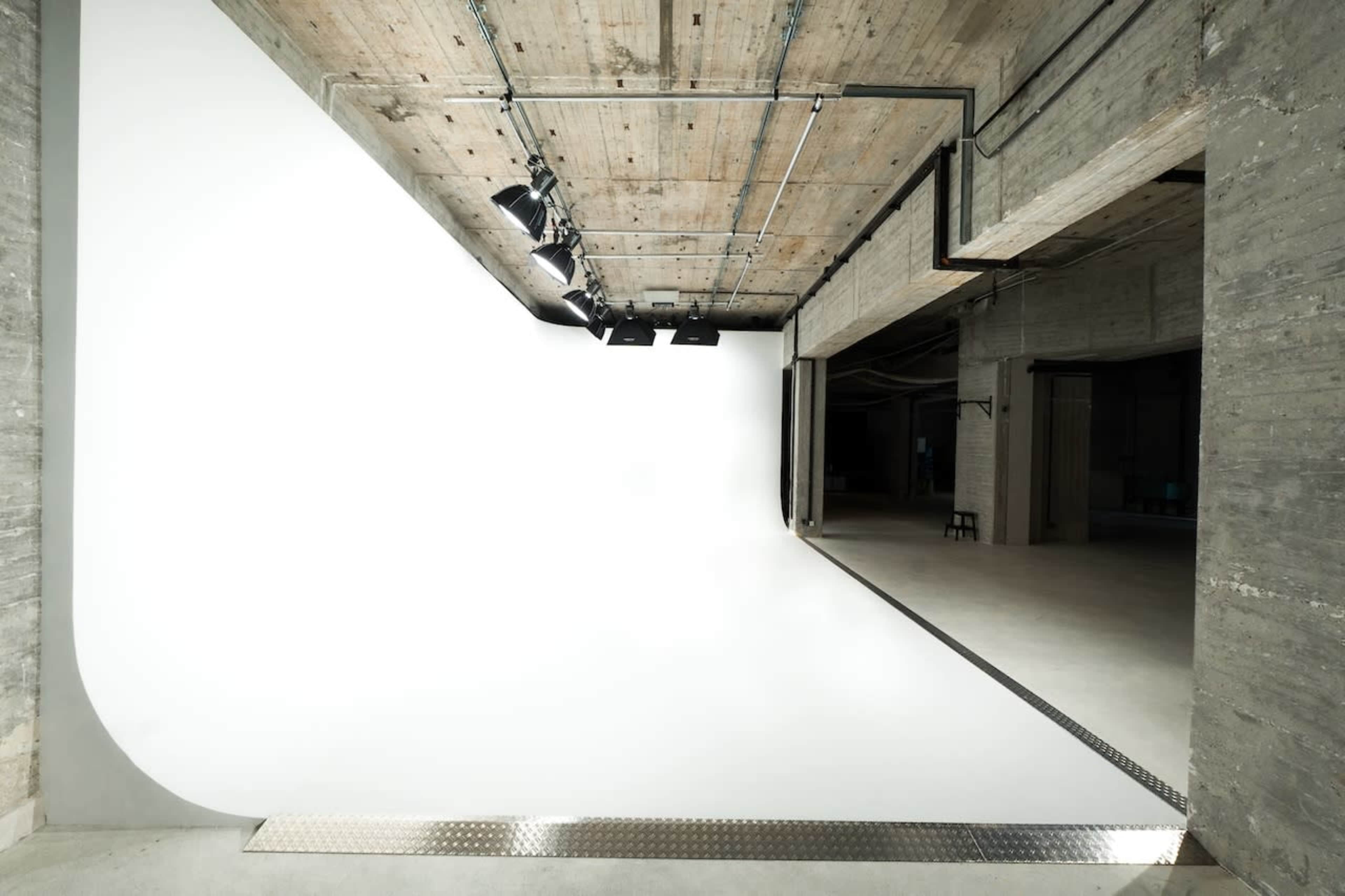 The image shows a spacious photography studio with a seamless white backdrop, overhead lighting, and concrete walls.