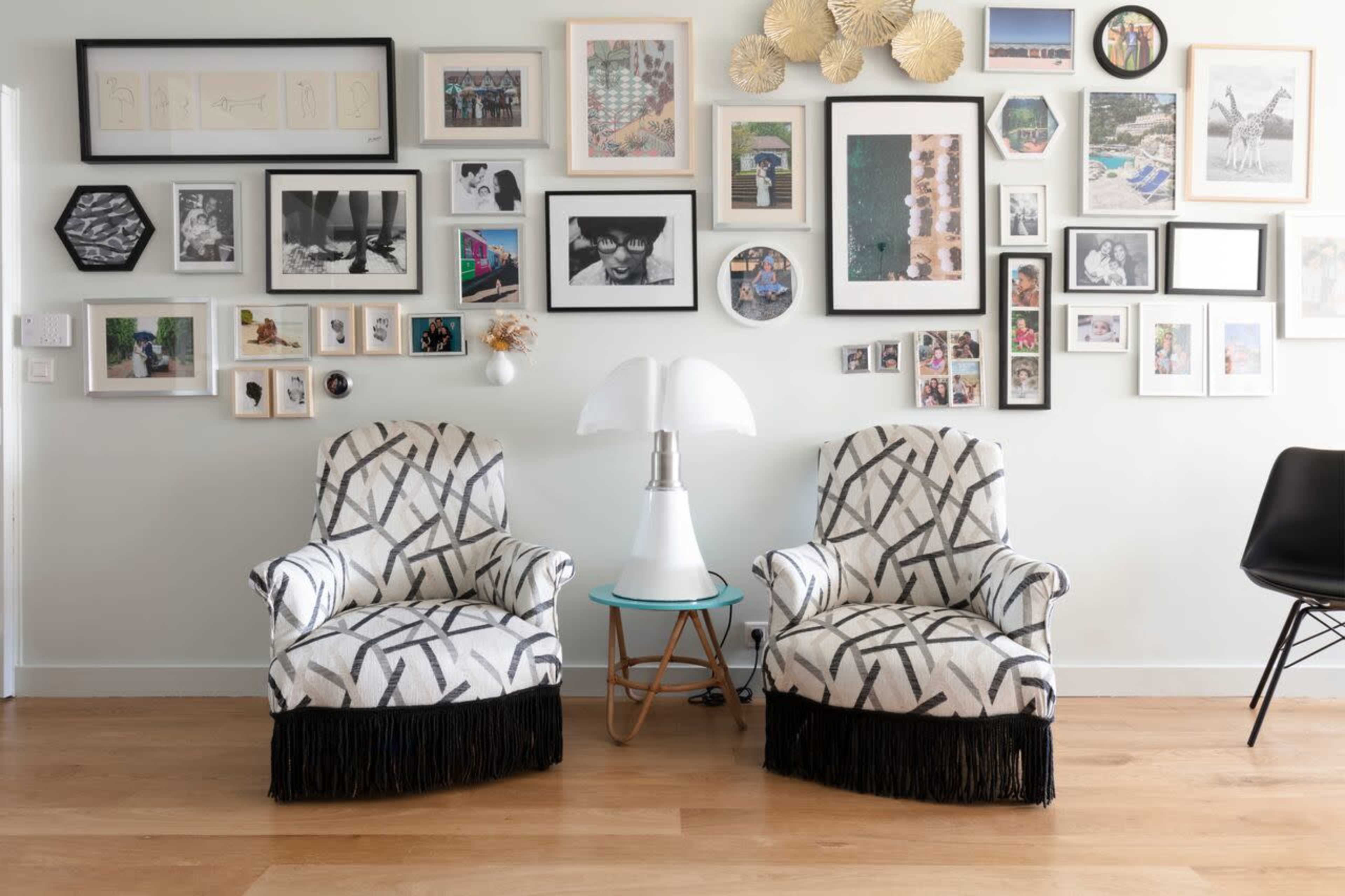 The image features two patterned armchairs with fringe placed in front of a gallery wall filled with framed photographs and artwork, alongside a small lamp and table.