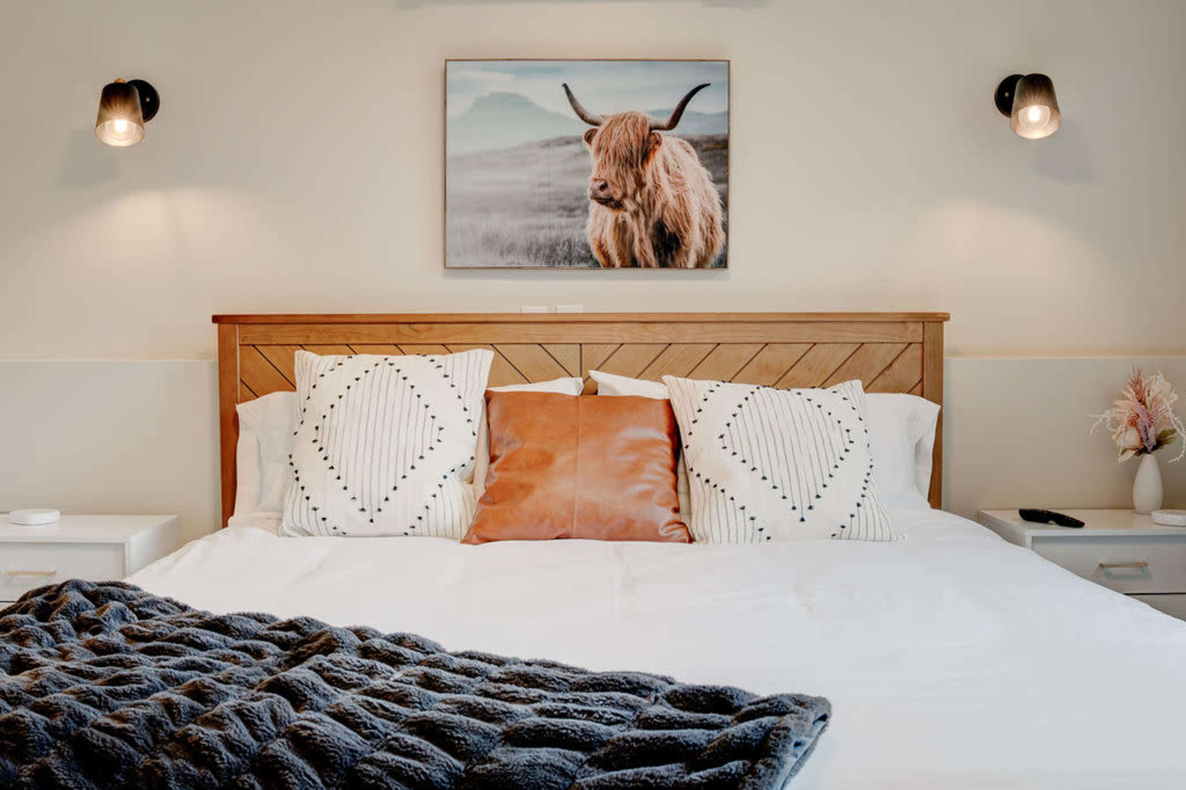 A neatly arranged bed with patterned pillows and a cozy blanket, adorned with a framed picture of a highland cow on the wall above.