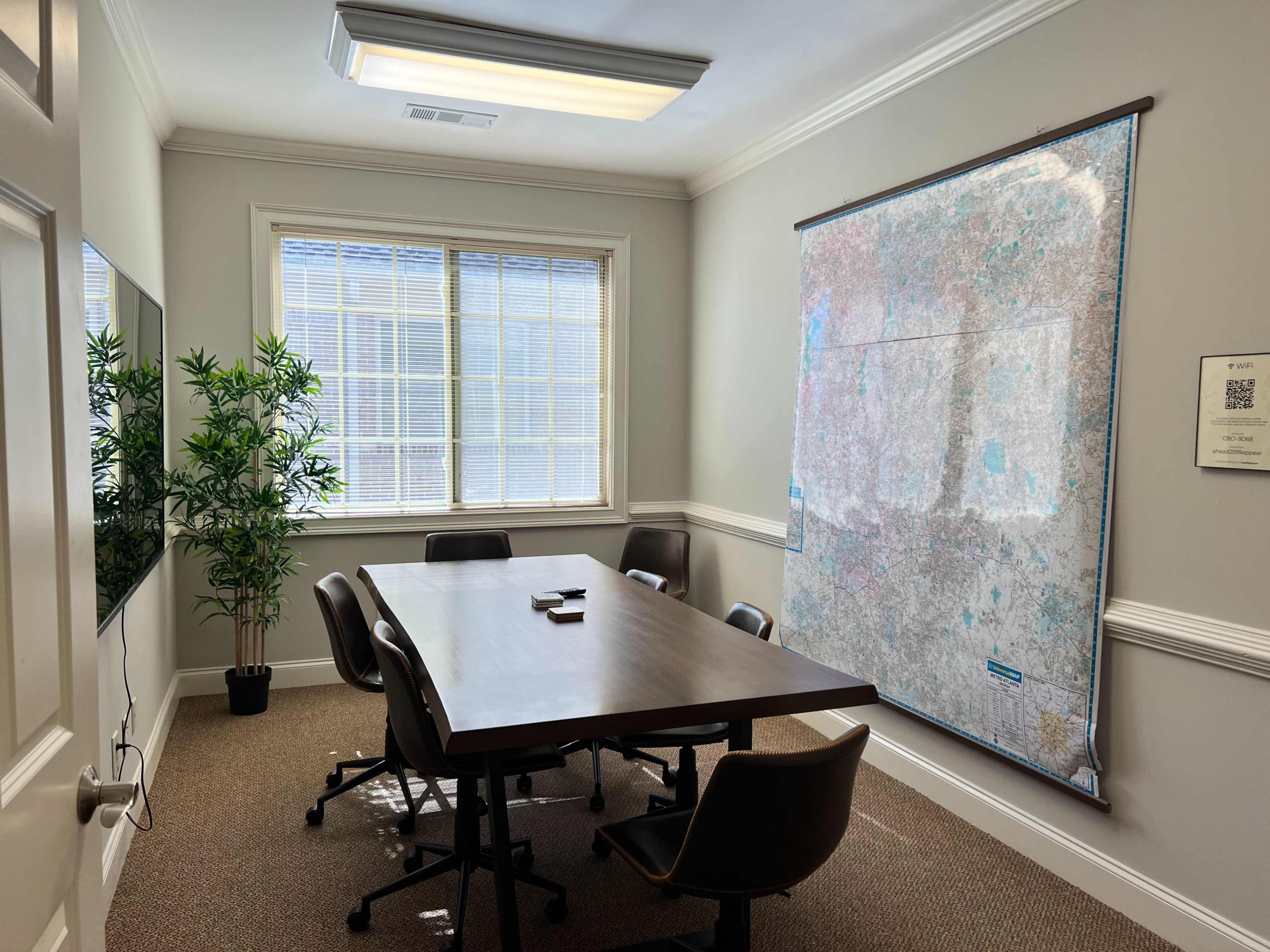 The image shows a conference room with a large wooden table surrounded by chairs, a wall map, a plant in the corner, and windows allowing natural light.