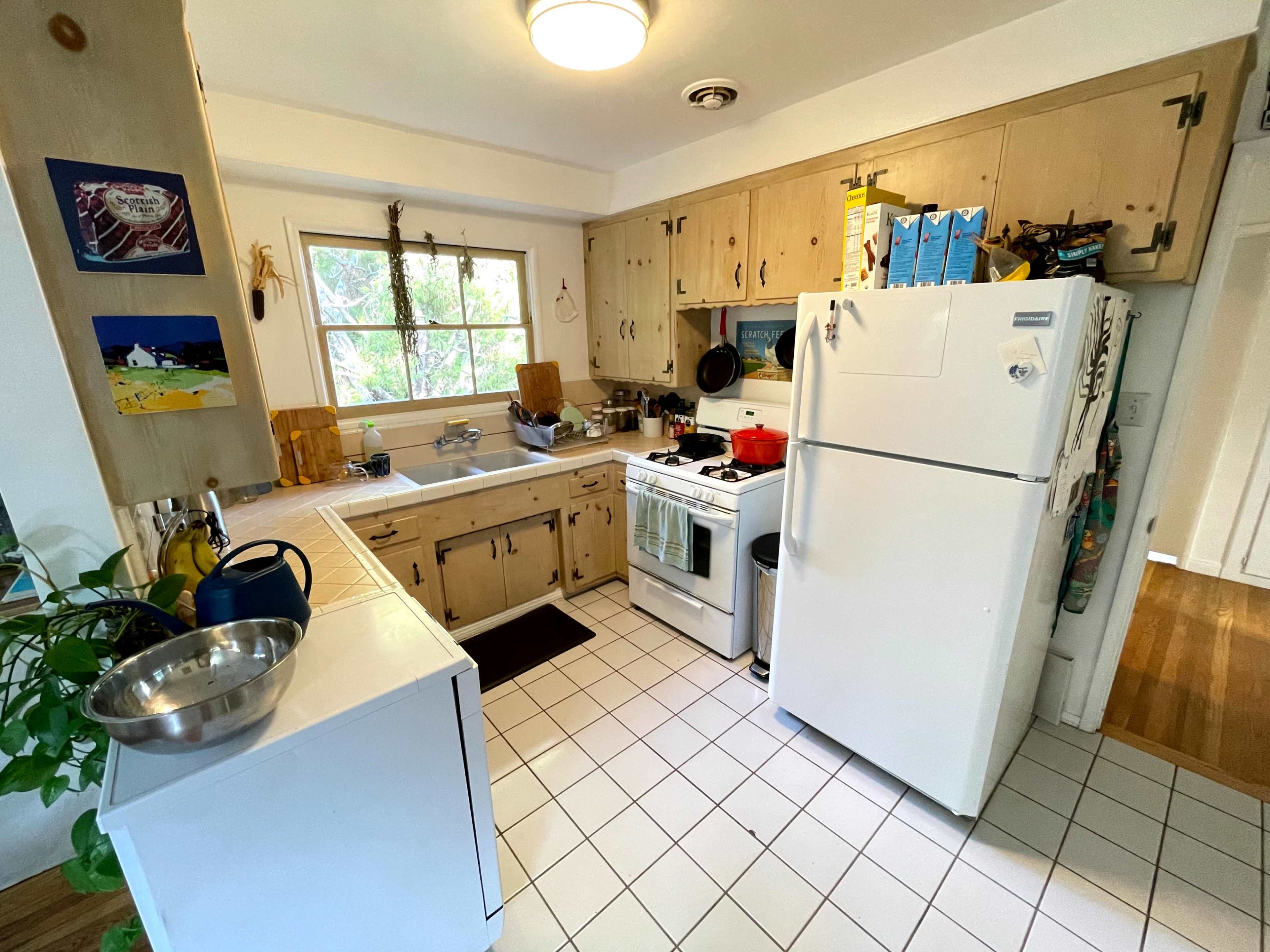 A small kitchen features a stove, refrigerator, and sink, surrounded by wooden cabinets and a window that provides natural light.