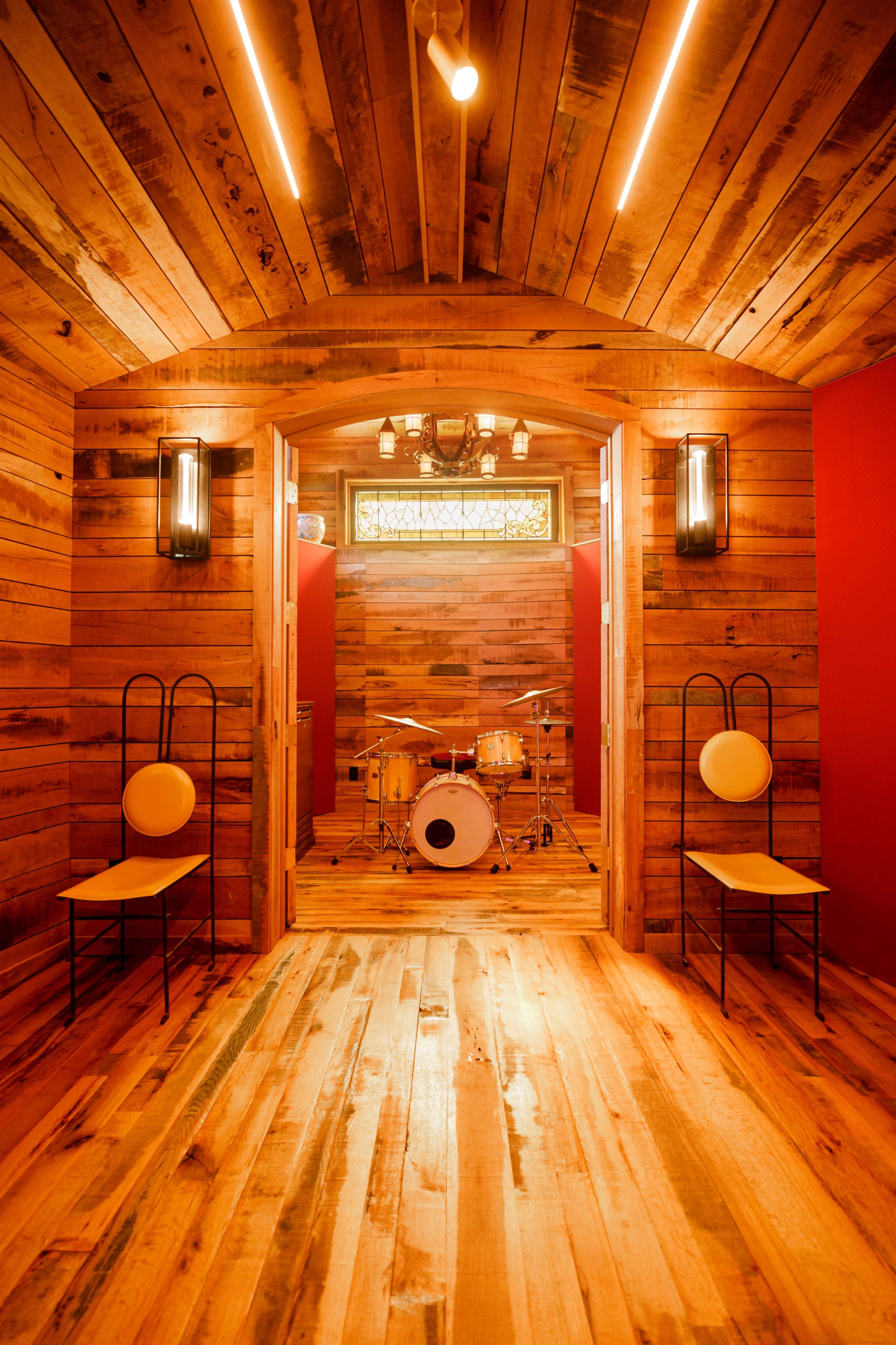 A wooden-paneled room featuring a drum set in the center, with two chairs positioned at the entrance.