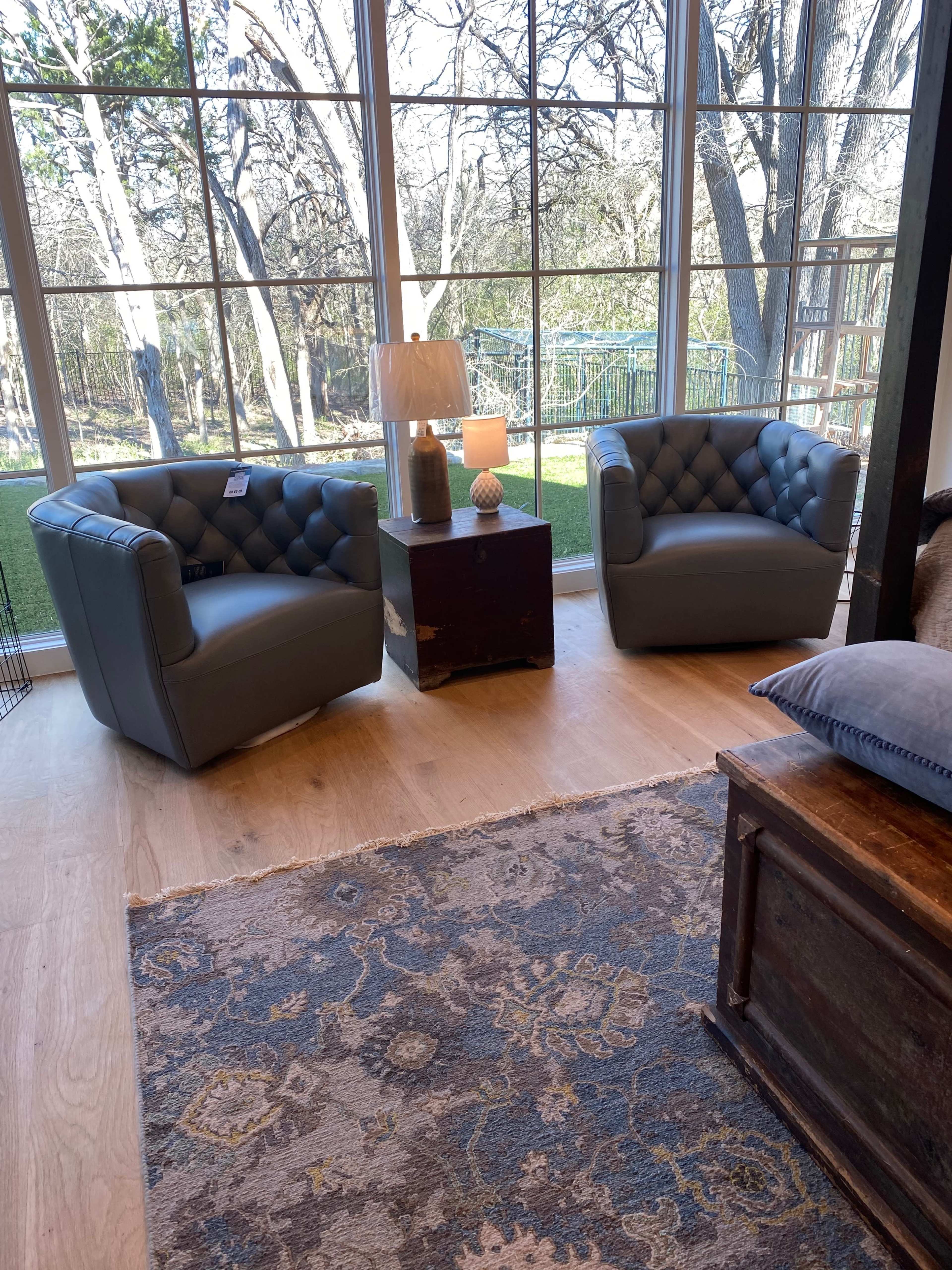 The image shows two gray tufted armchairs positioned near a window, alongside a small table with a lamp and decorative items.