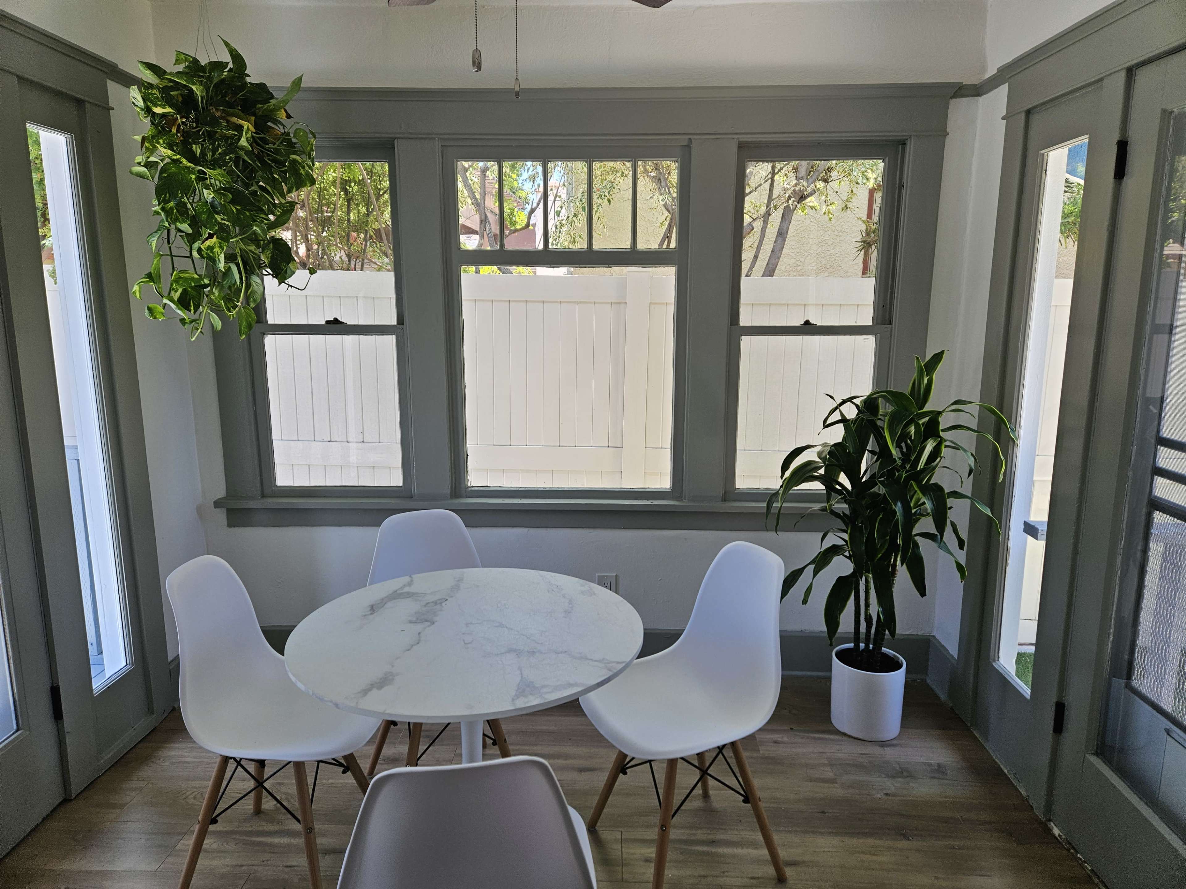A bright dining area features a round marble table surrounded by white chairs, with large windows providing natural light and potted plants in the corners.