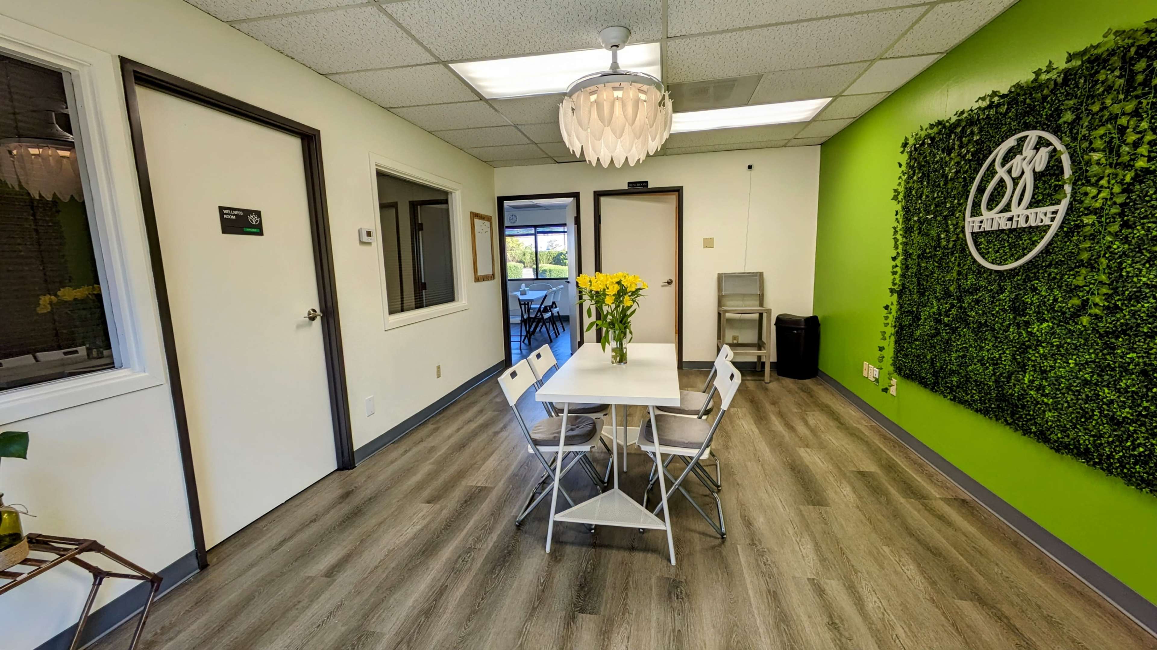 A bright, modern waiting room features a white table surrounded by four folding chairs, green accent walls, and potted plants.