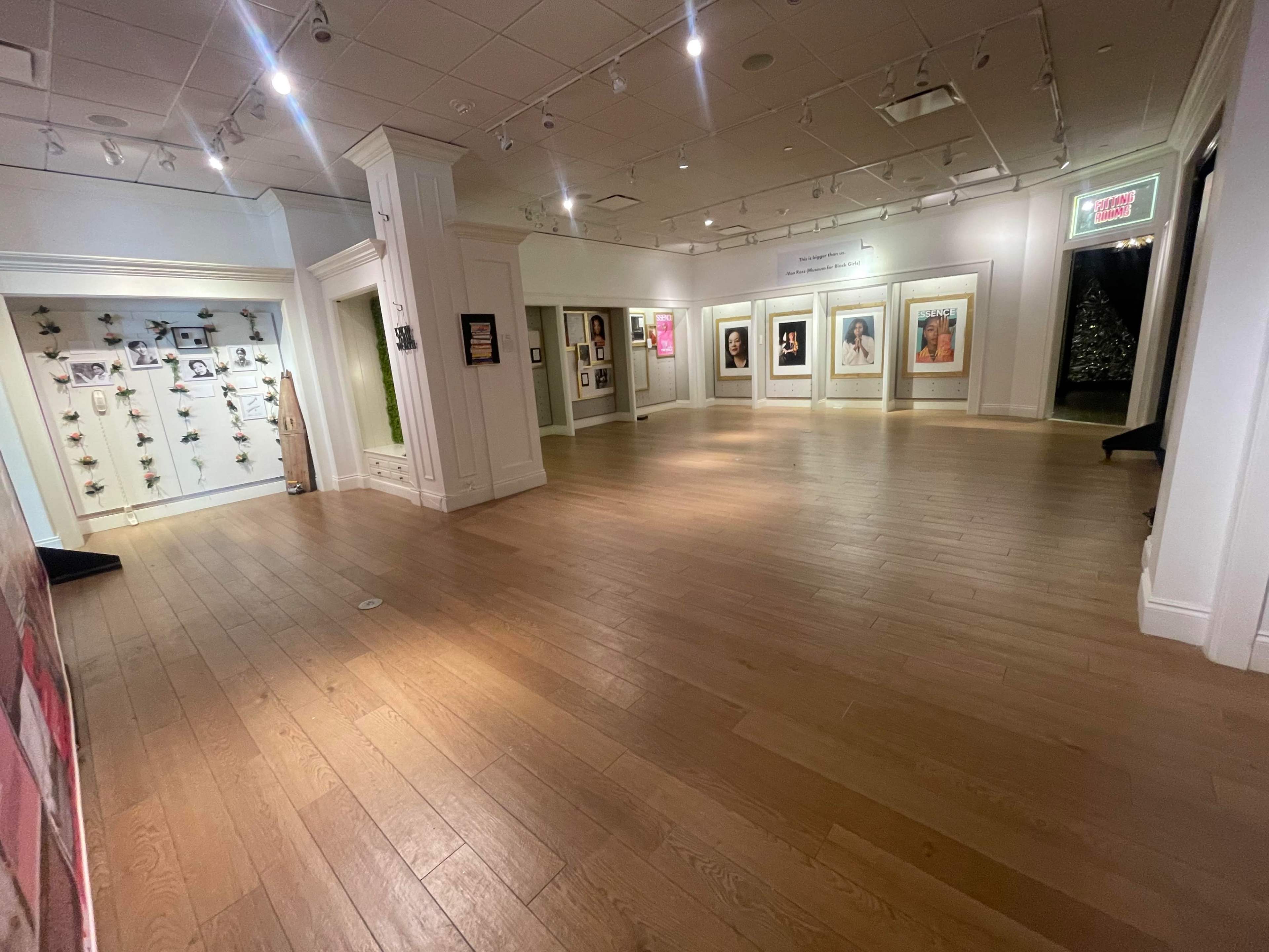 The image shows a spacious, well-lit art gallery with wooden flooring and framed artworks displayed on the walls.