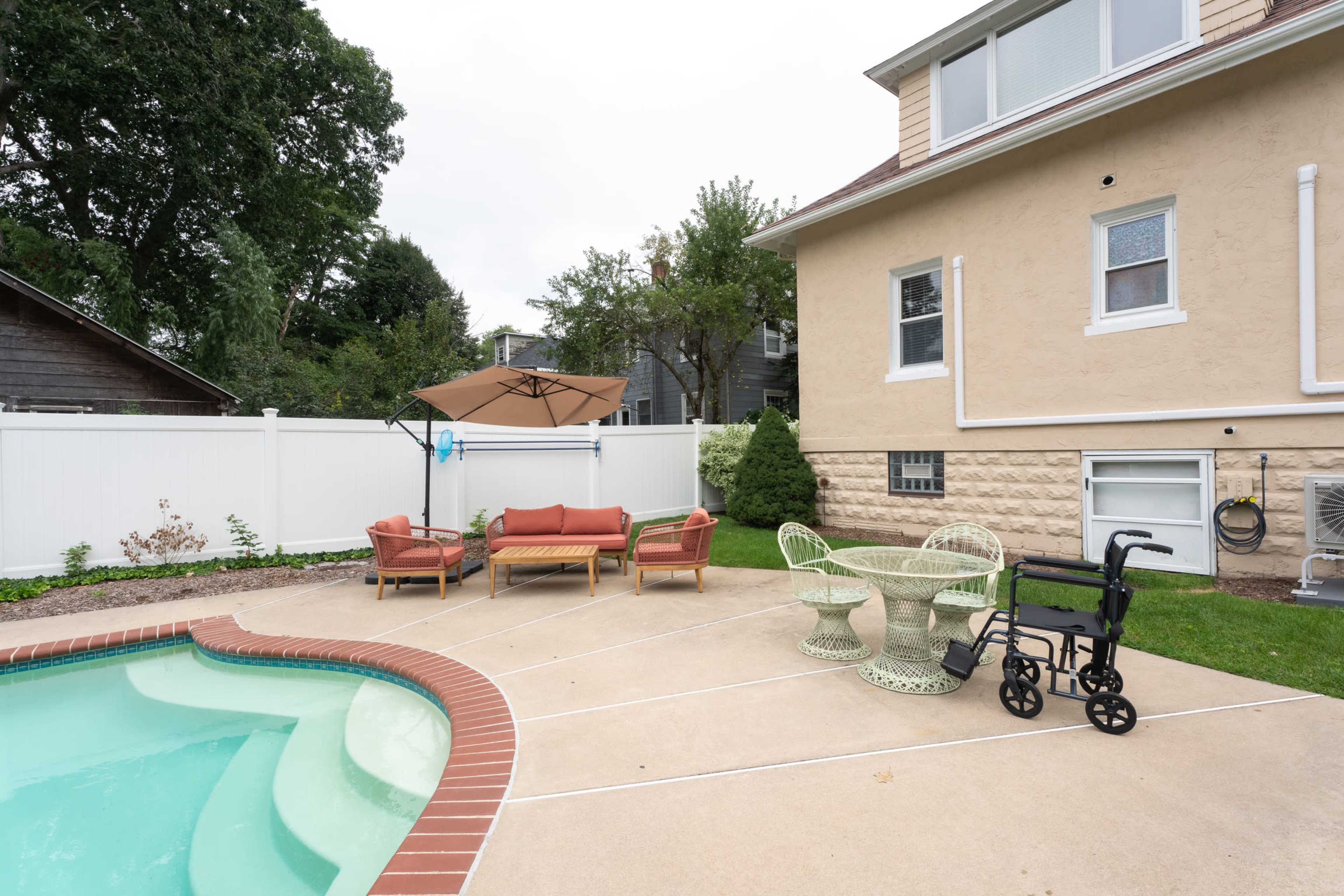 The image shows a backyard with a swimming pool, a seating area featuring brown couches and a green table, and a wheelchair beside the pool.