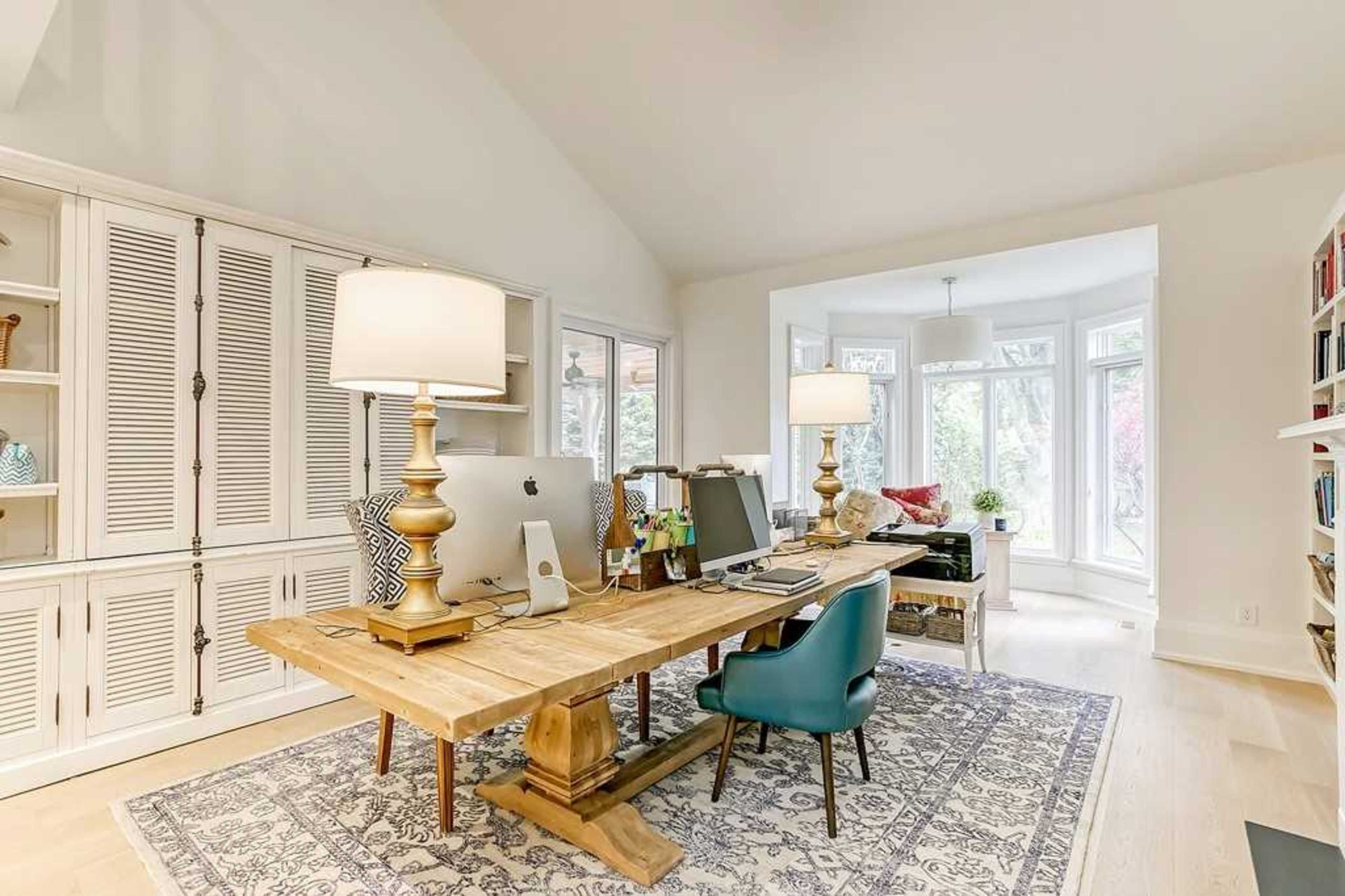 A spacious home office features a wooden table with a computer, two lamps, and a blue chair, surrounded by built-in cabinets and large windows overlooking a garden.
