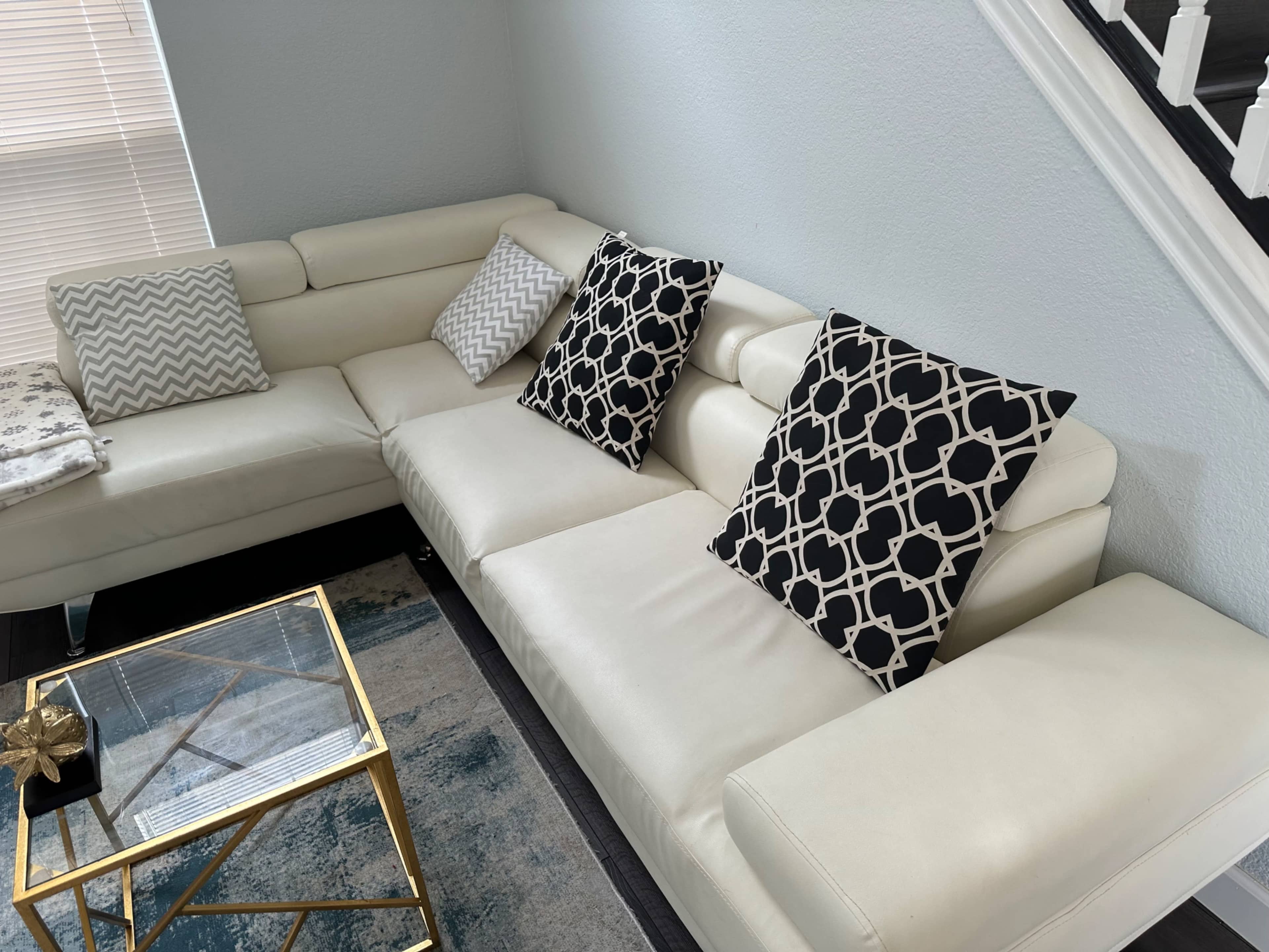 A white sectional sofa with patterned pillows sits beside a glass coffee table on a rug in a well-lit room.