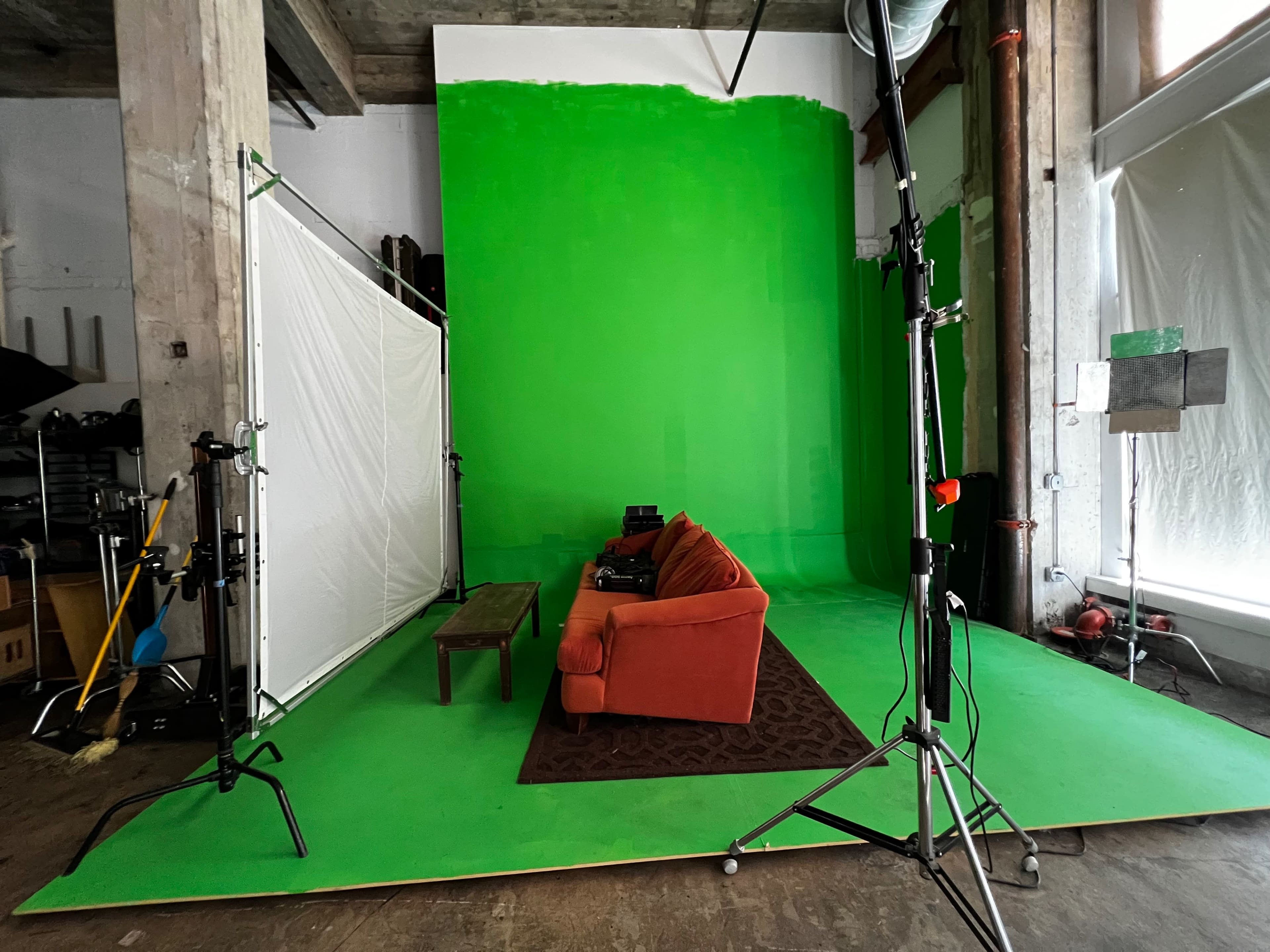The image shows a green screen studio setup featuring an orange couch, a coffee table, and a patterned rug on a green floor.