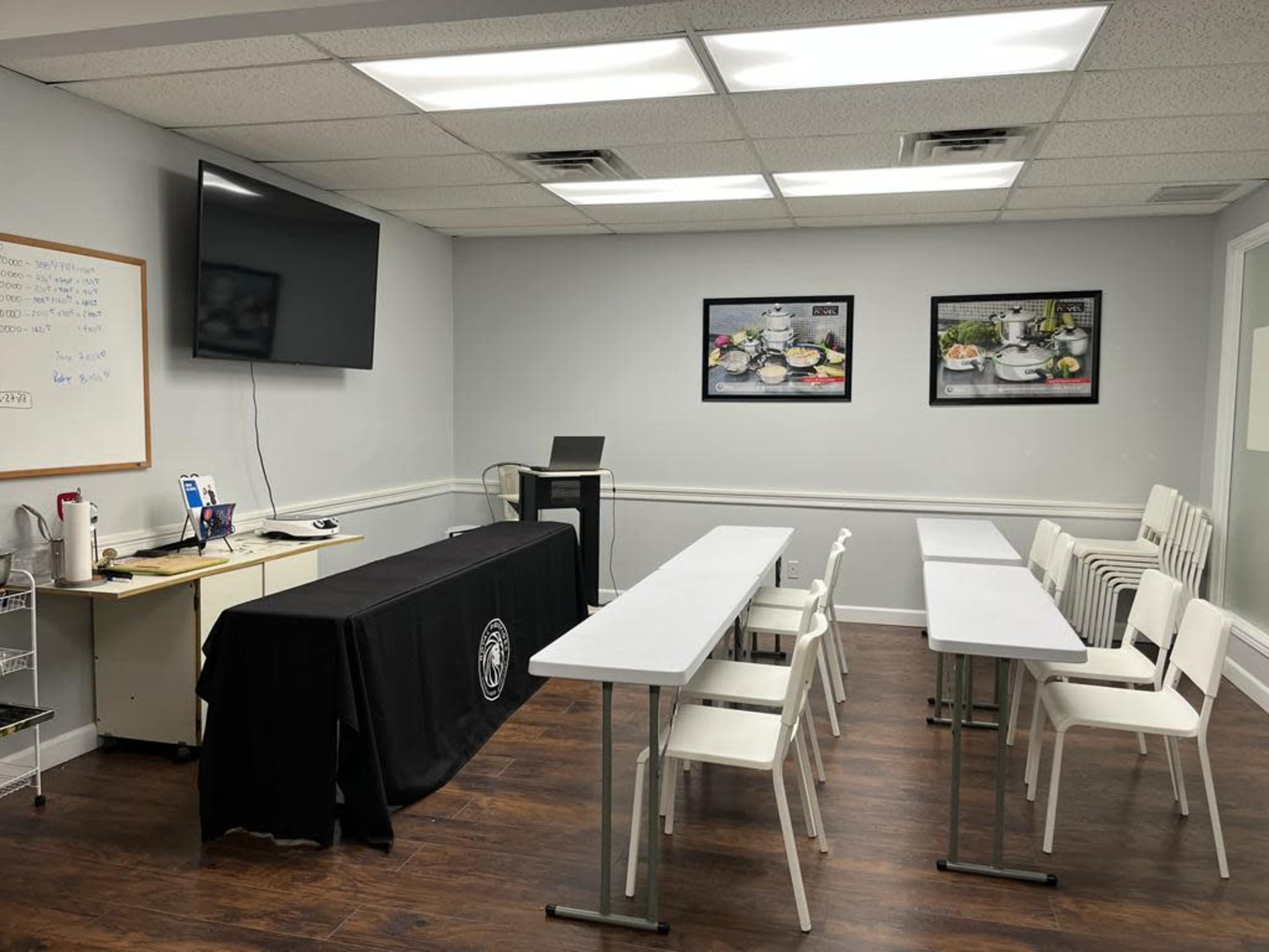 A classroom setting with a long table covered by a black tablecloth, several white chairs arranged around tables, and a large screen mounted on the wall.