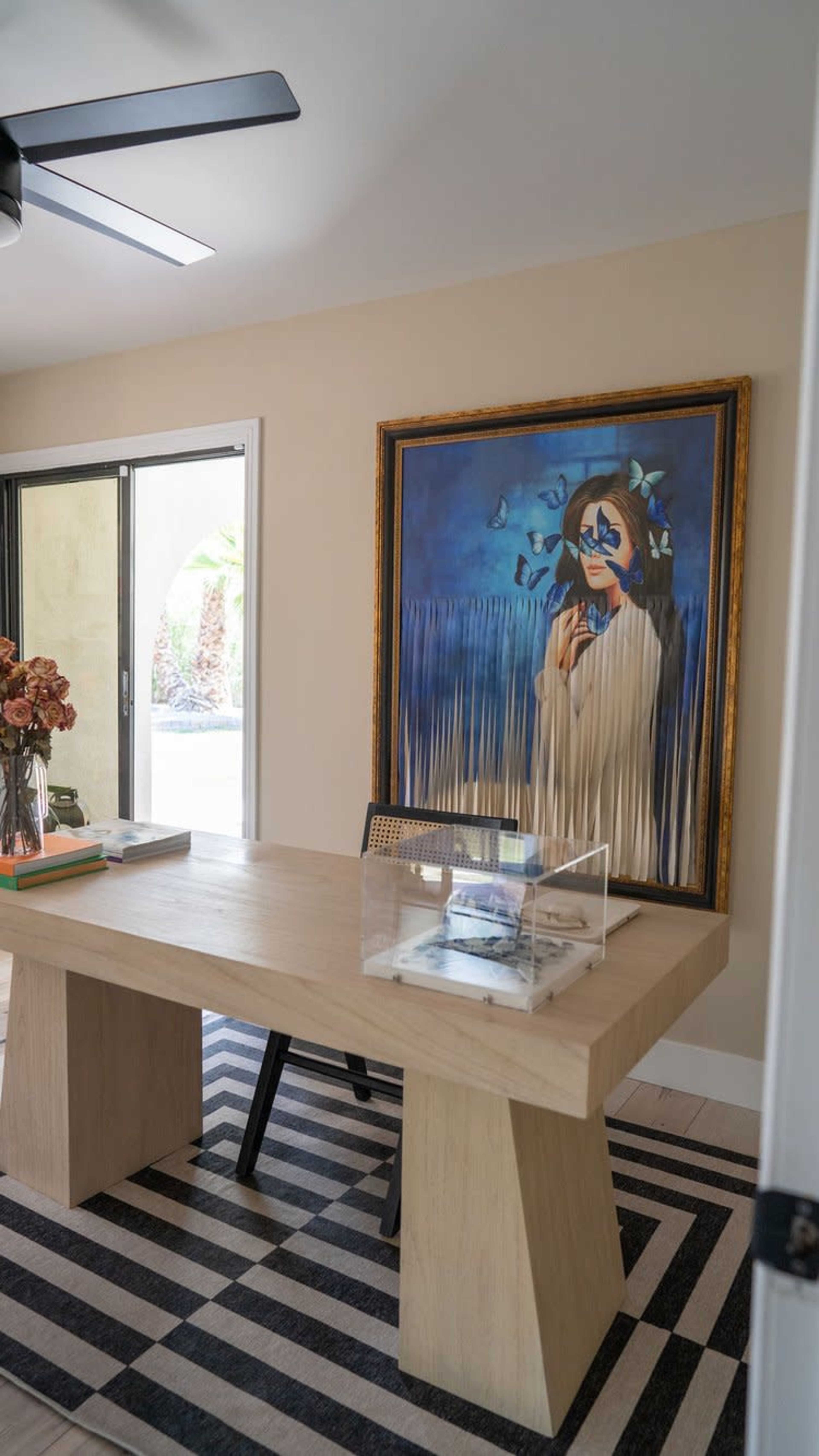 The image shows a modern office space with a large wooden table, a framed painting of a woman with blue butterflies, and a striped rug underneath.