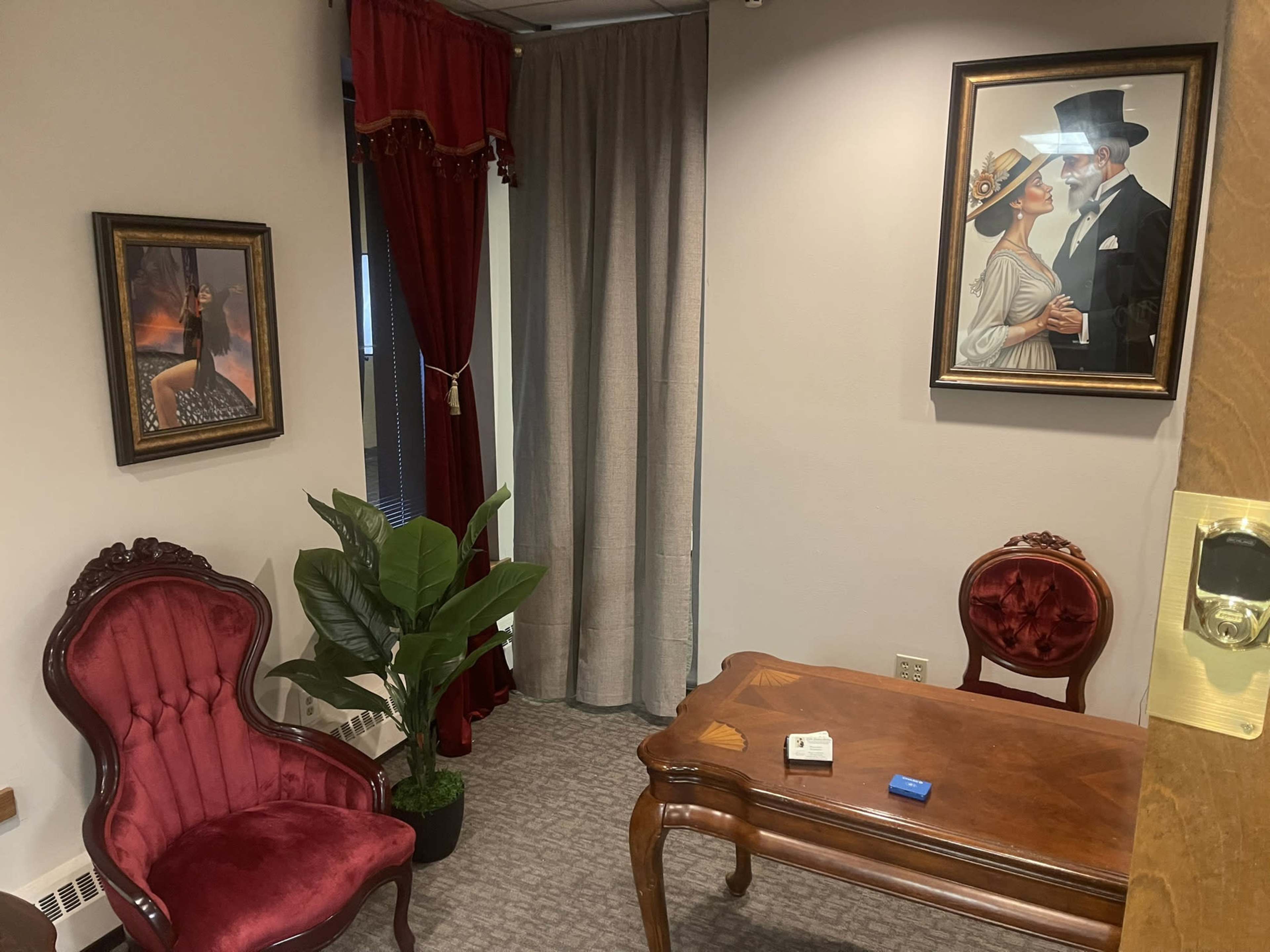 The image shows a small office space furnished with a wooden table, two red velvet chairs, a potted plant, and framed artwork on the walls.