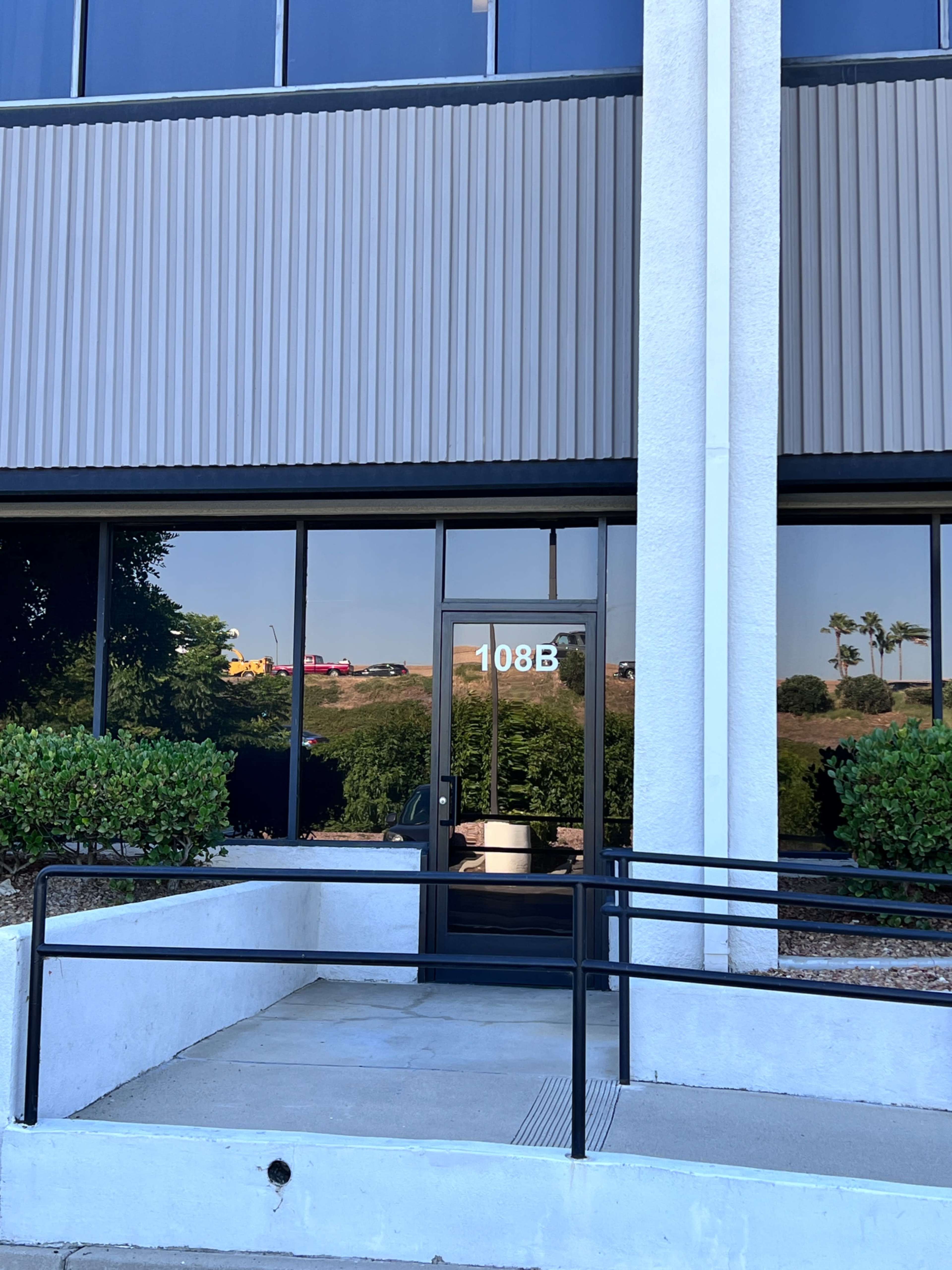 The entrance of a commercial building labeled "108B" with a ramp and landscaped greenery near the door.