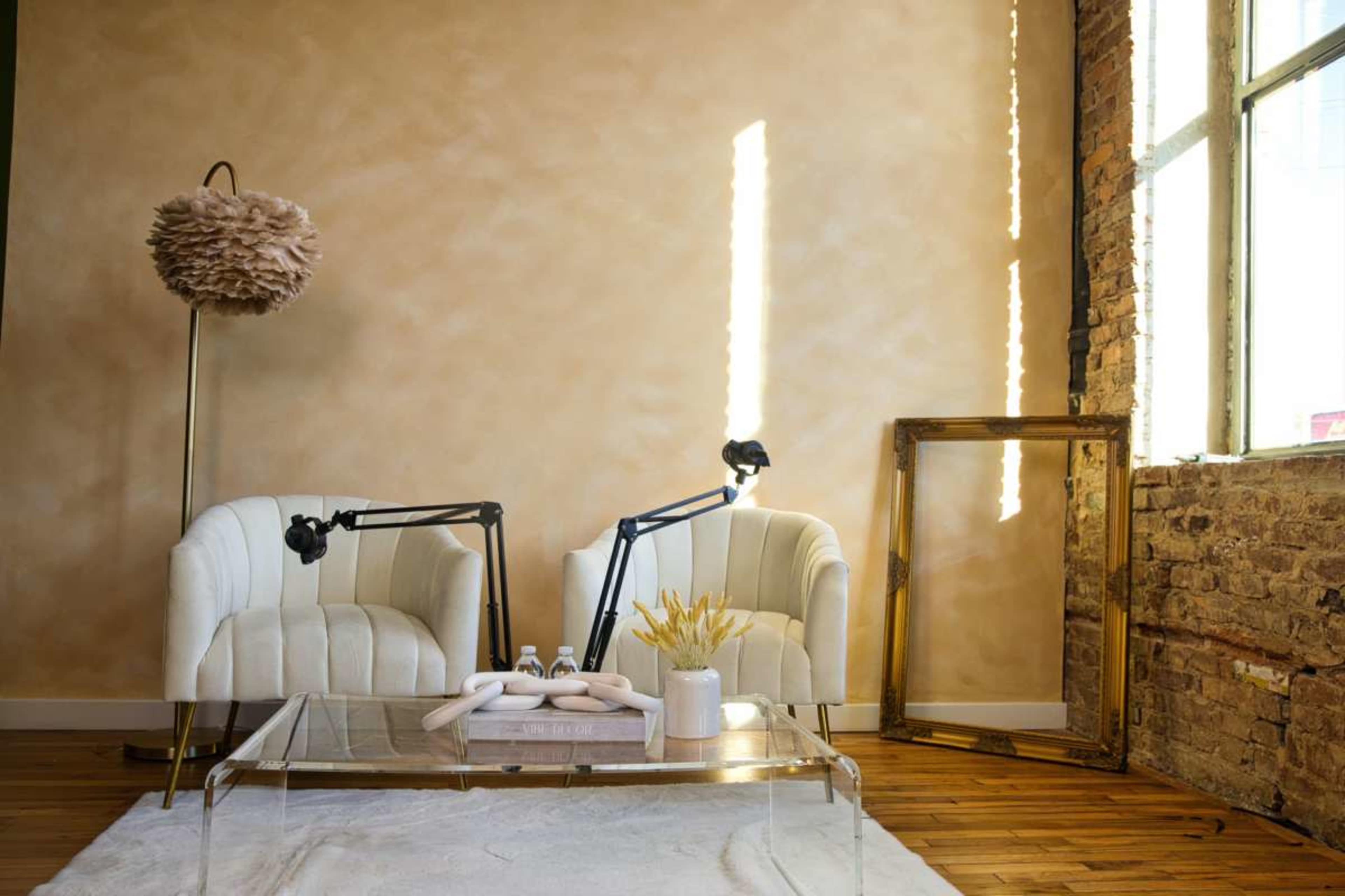 The image features a stylish interior with two white chairs, a glass coffee table, and a standing lamp, set against a softly colored wall and exposed brick.
