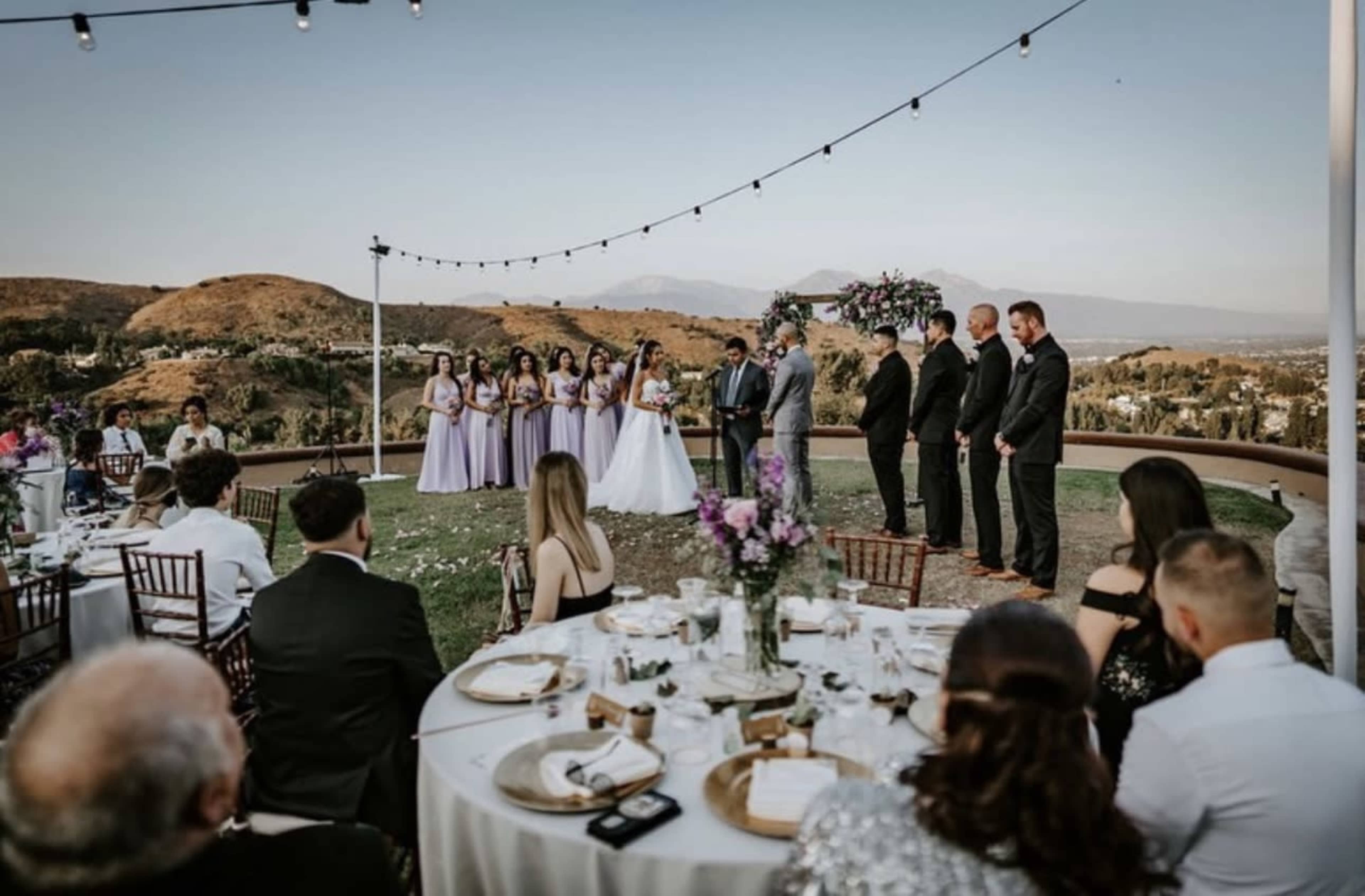 A wedding ceremony takes place outdoors with guests seated at decorated tables, and the bridal party standing under string lights and floral arrangements.