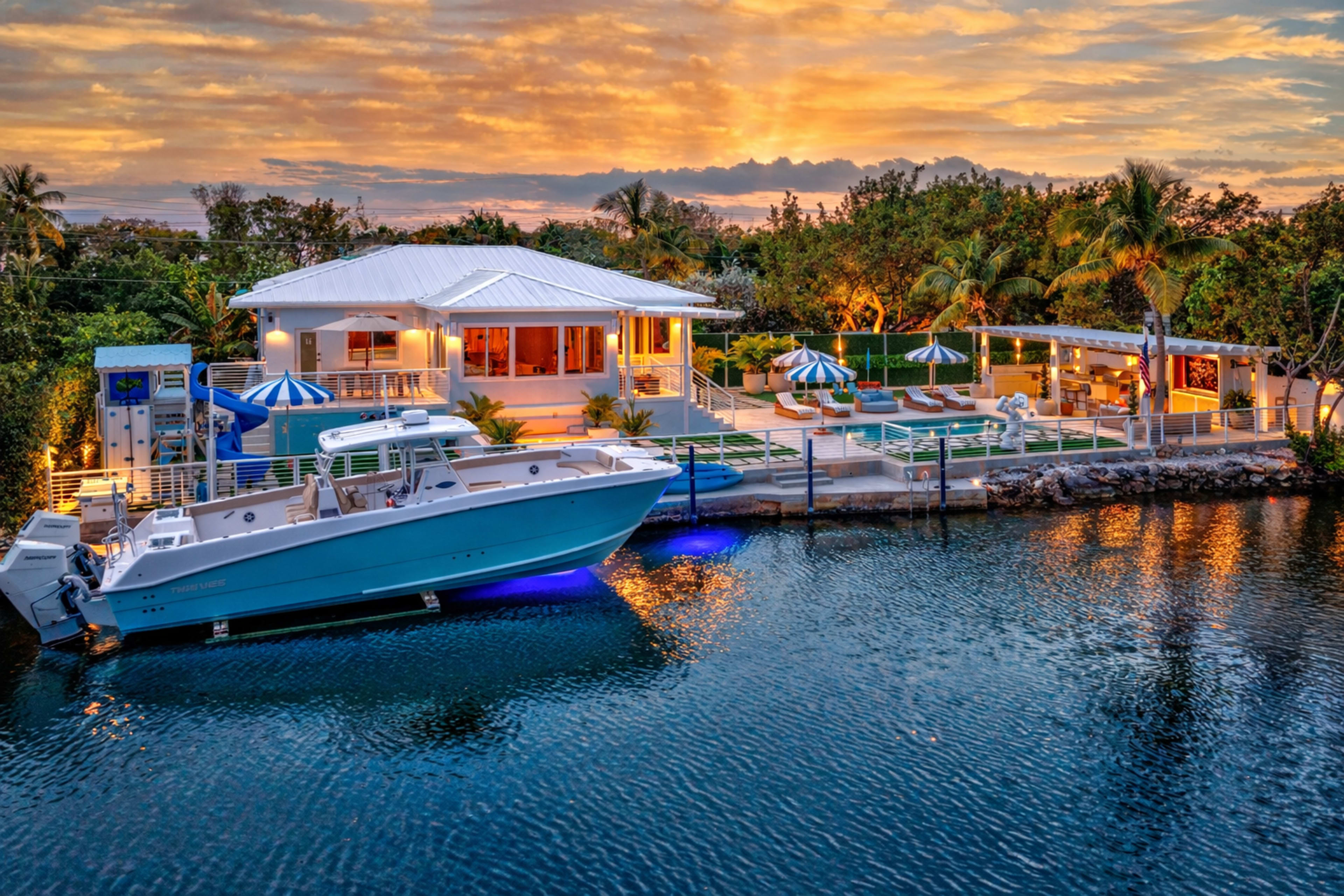 The image shows a modern waterfront property with a boat docked in front, surrounded by palm trees, a swimming pool, and large umbrellas, all illuminated by a vibrant sunset.