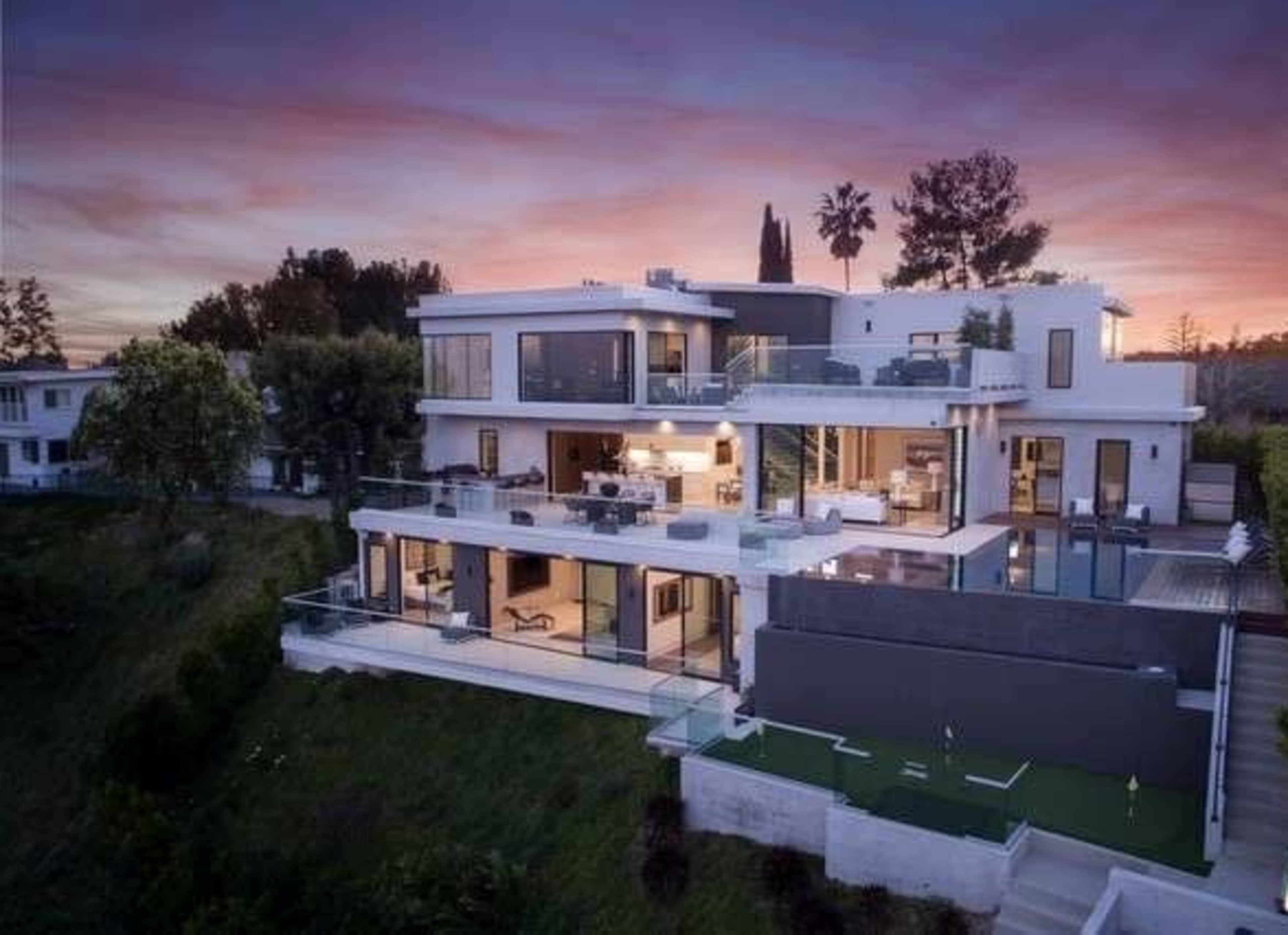 A modern multi-level house with large glass windows and a terrace, situated on a hillside at sunset.