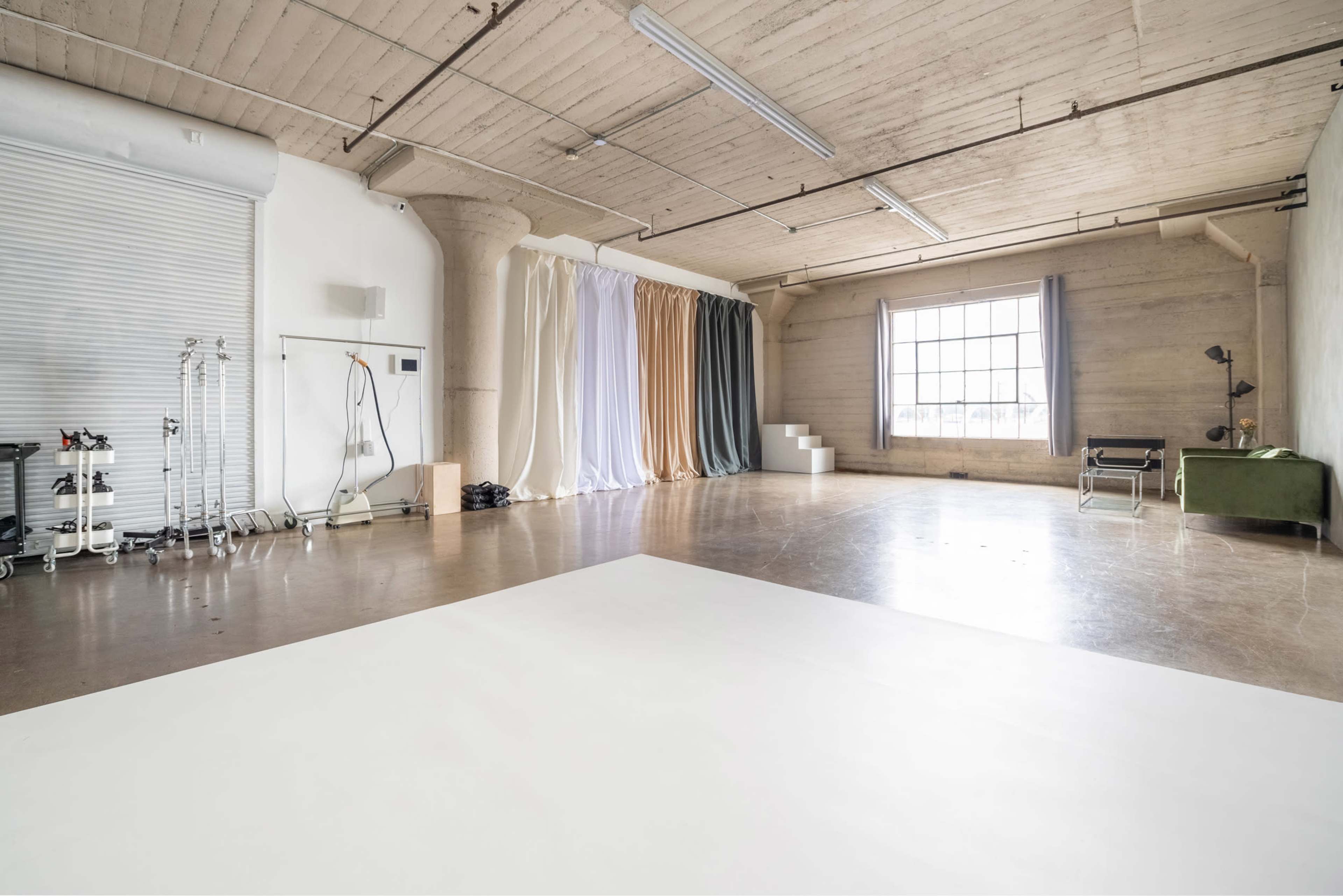 The image shows a spacious, minimalist photography studio with fabric backdrops arranged along one wall and a large window providing natural light.