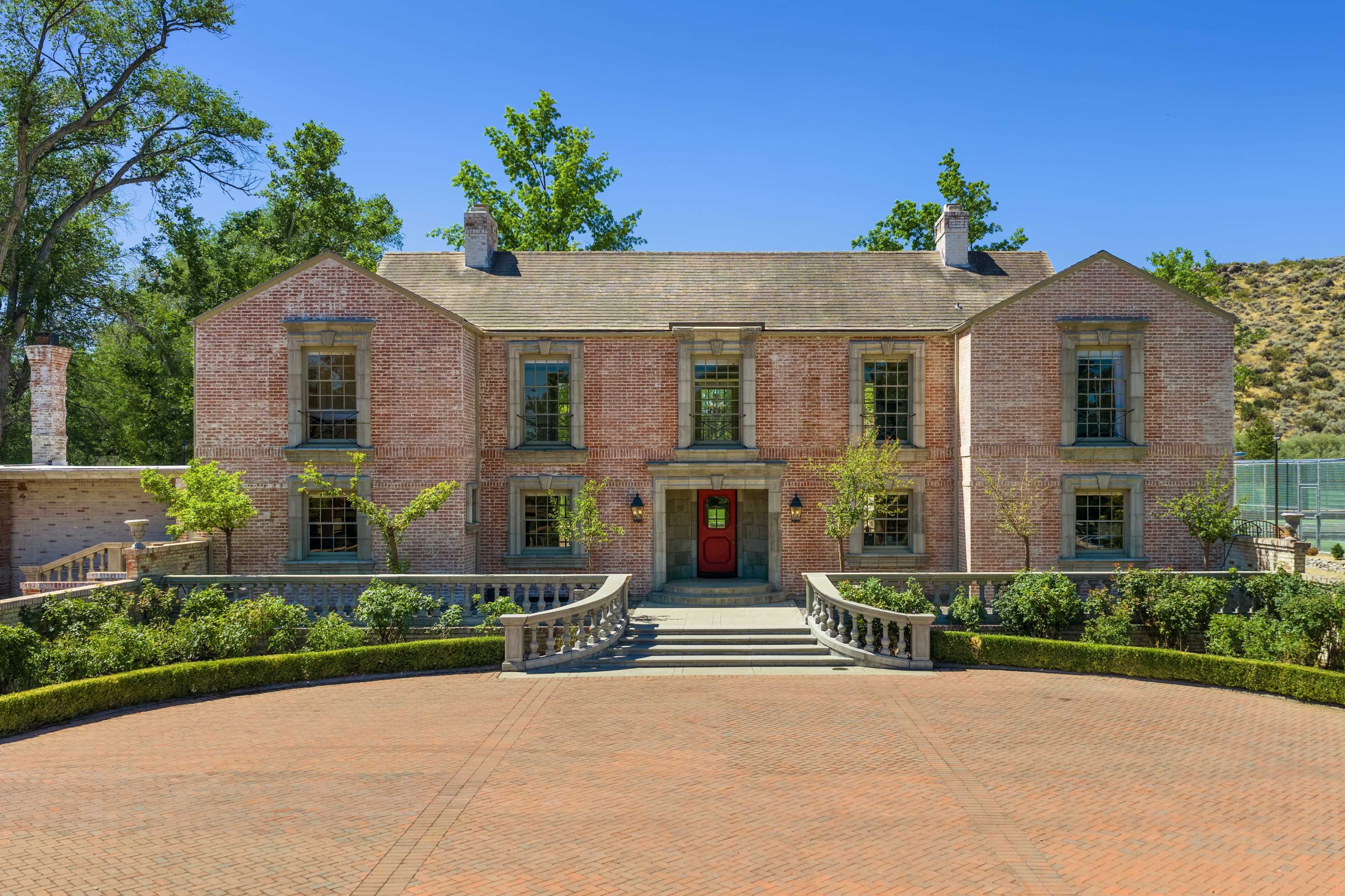 The image shows a large, two-story brick house with a symmetrical facade, featuring a central red door, surrounded by landscaped gardens and a circular driveway.