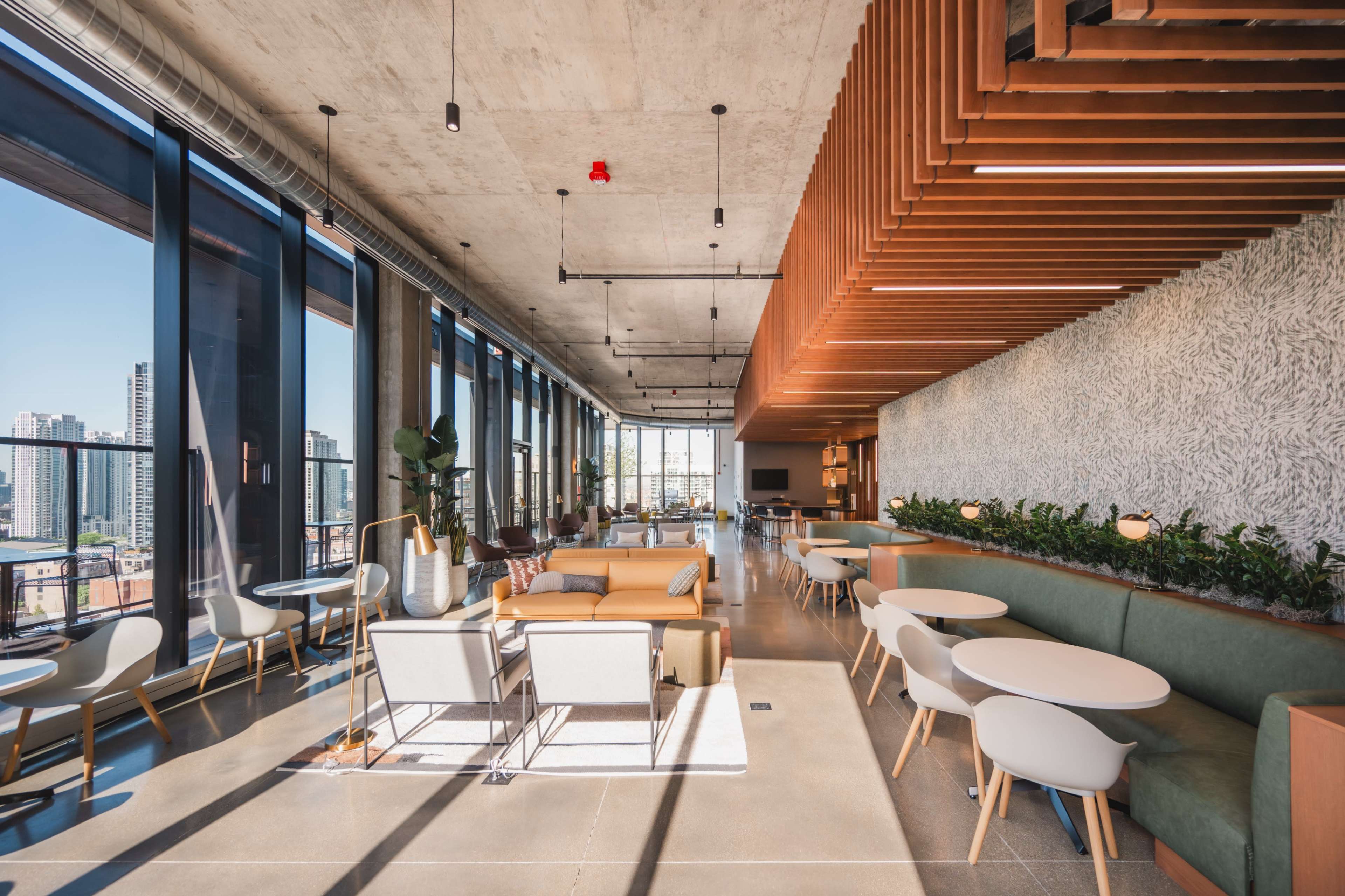 The image shows a modern office lounge with large windows, wooden ceiling beams, and various seating arrangements, including sofas and round tables.