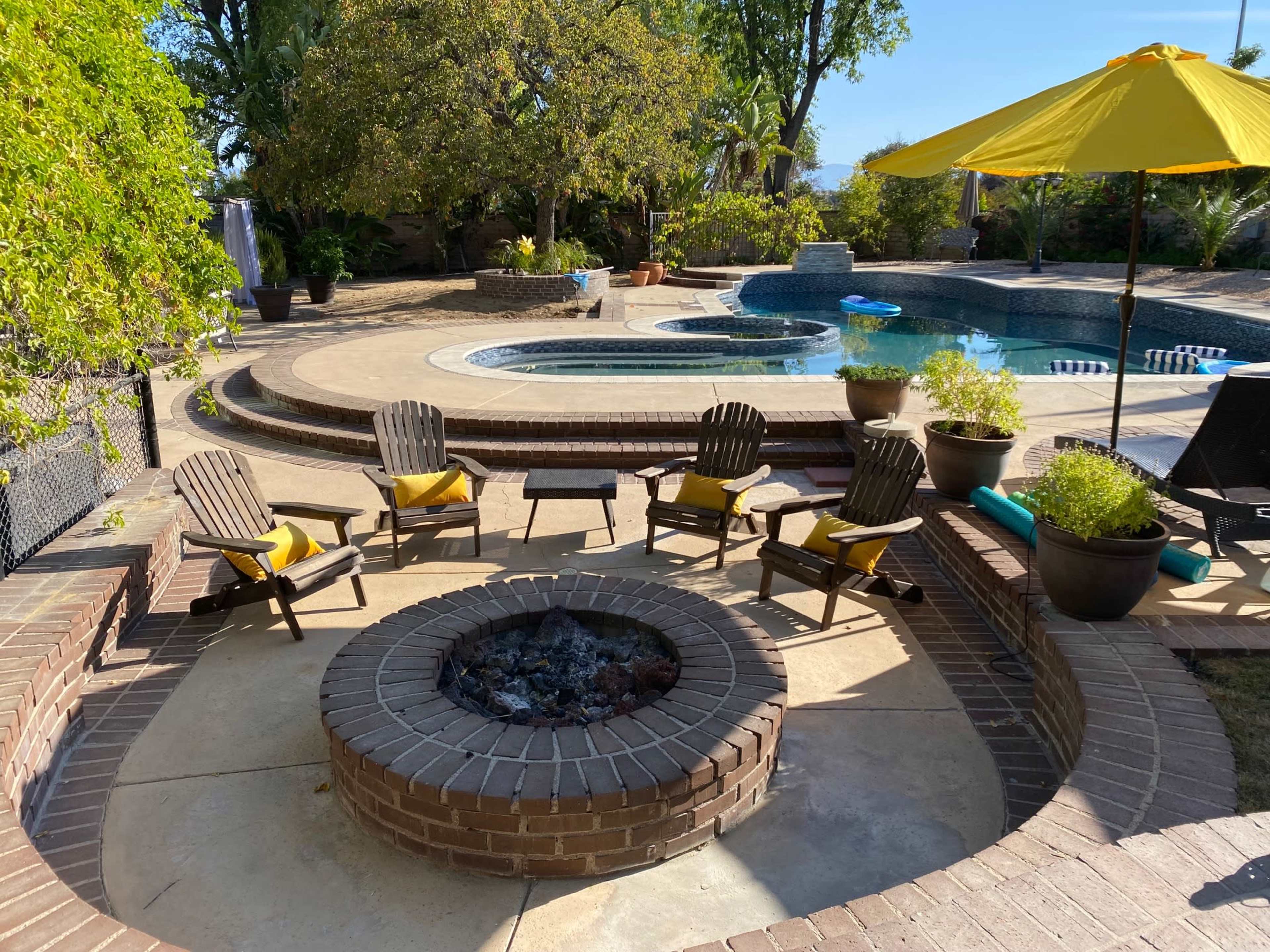 The image shows a backyard with a fire pit surrounded by wooden chairs, a swimming pool, and a yellow umbrella.