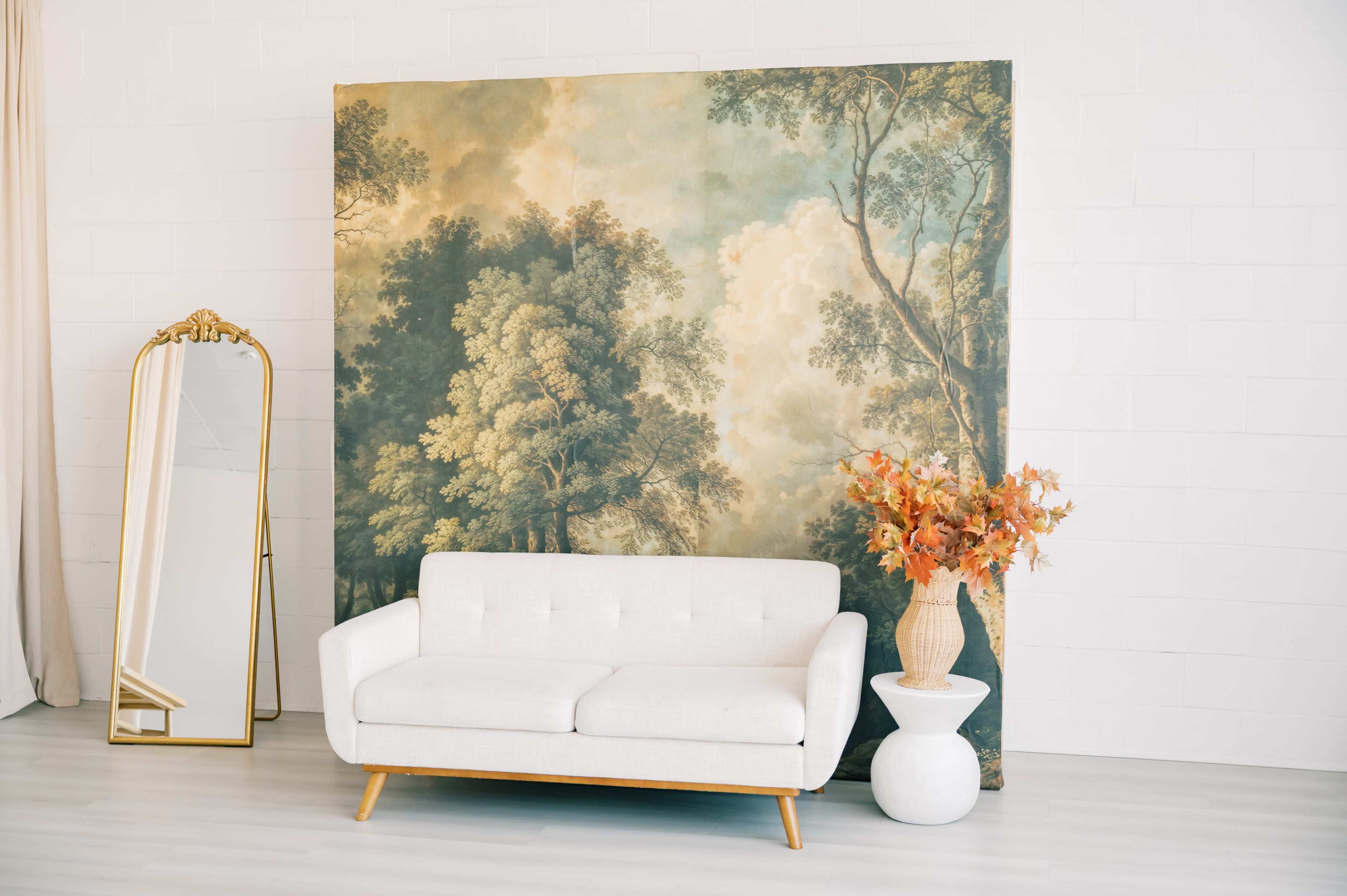 A white sofa is positioned in front of a large mural depicting trees and a cloudy sky, with a mirror and a vase of autumn flowers nearby.