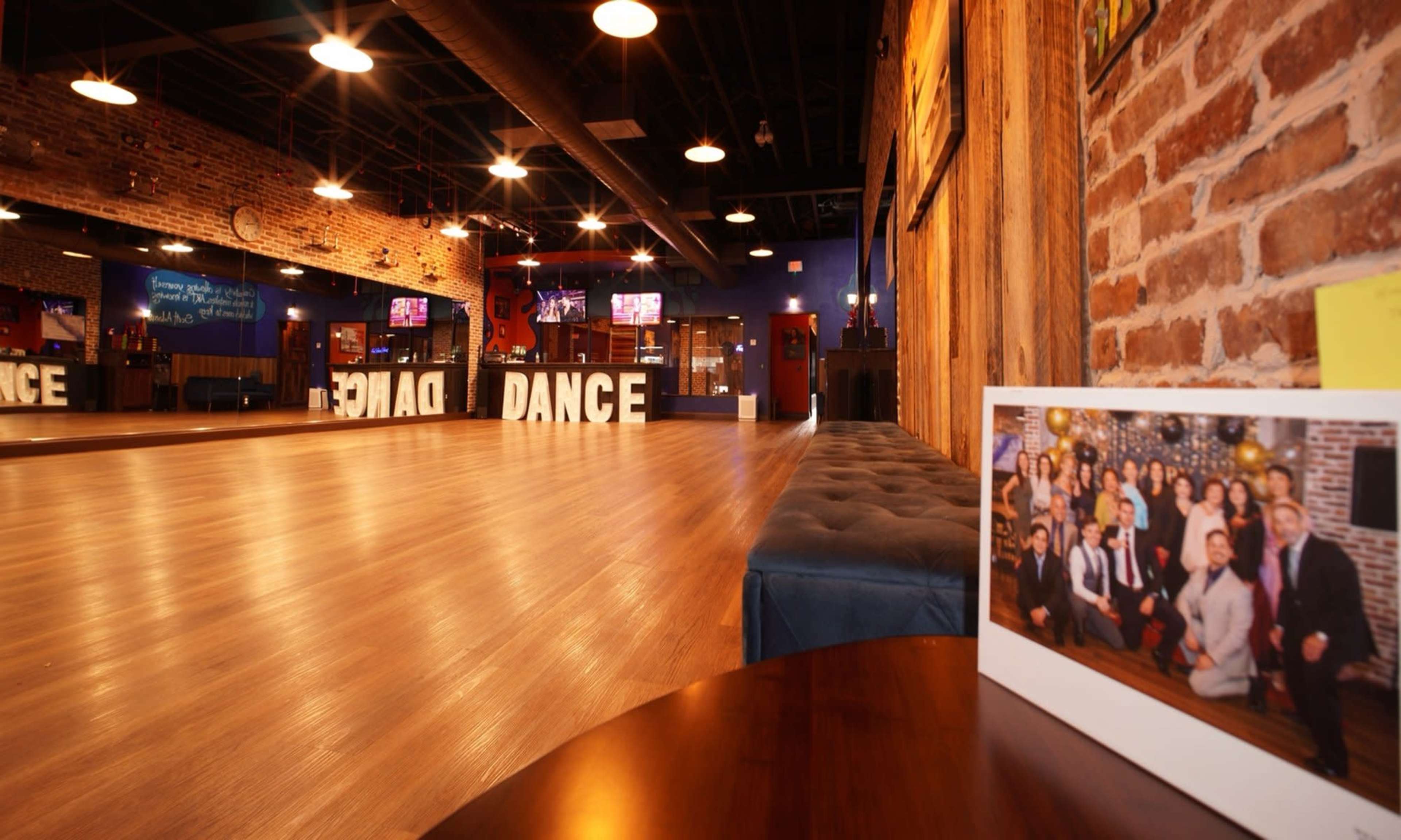 The image shows a spacious dance studio with wooden floors, exposed brick walls, and a large "DANCE" sign prominently displayed in the background.