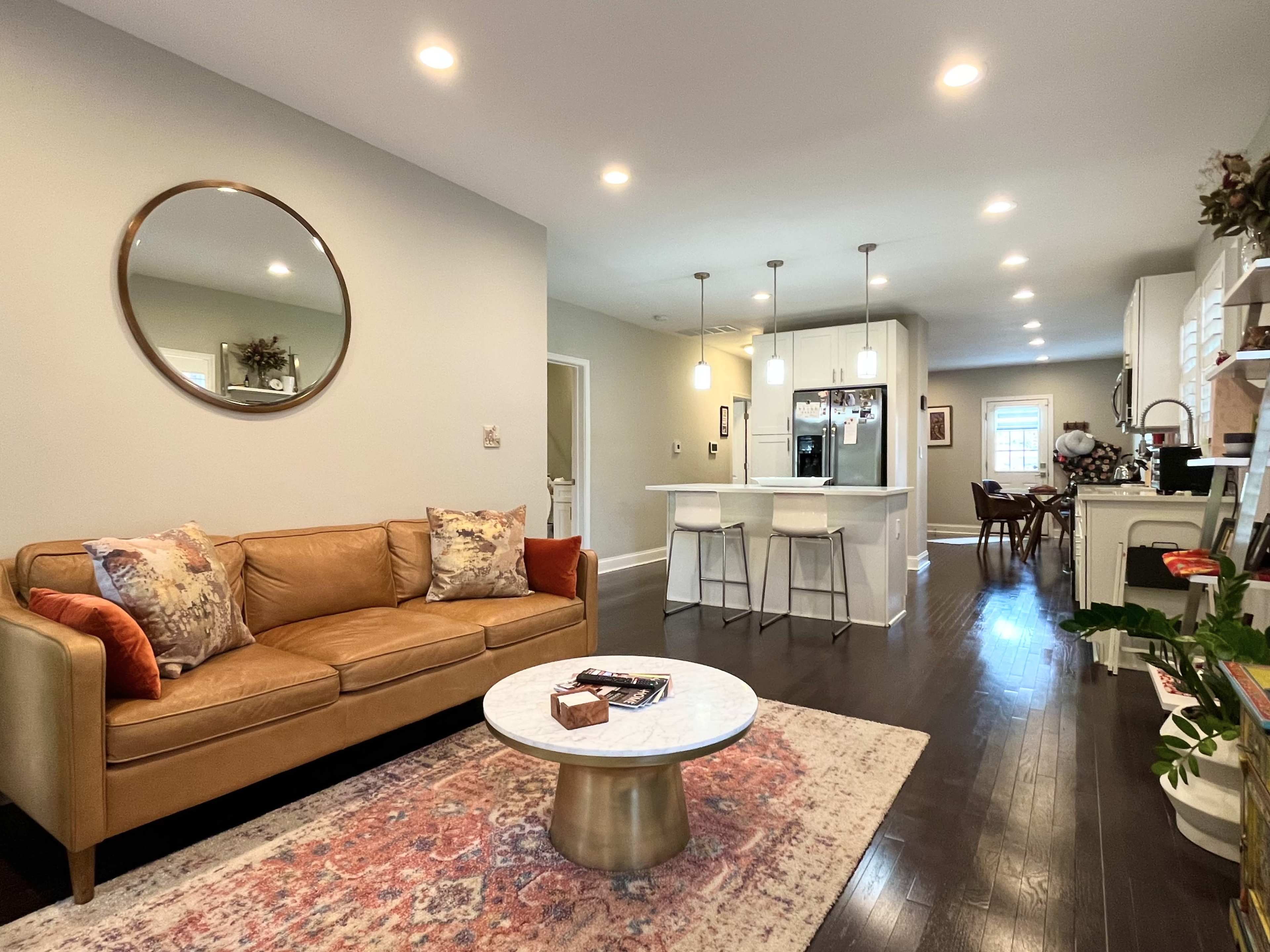 A living room with a brown sofa, decorative pillows, a round mirror on the wall, and a coffee table, leading into a kitchen area that features barstools and bright lighting.