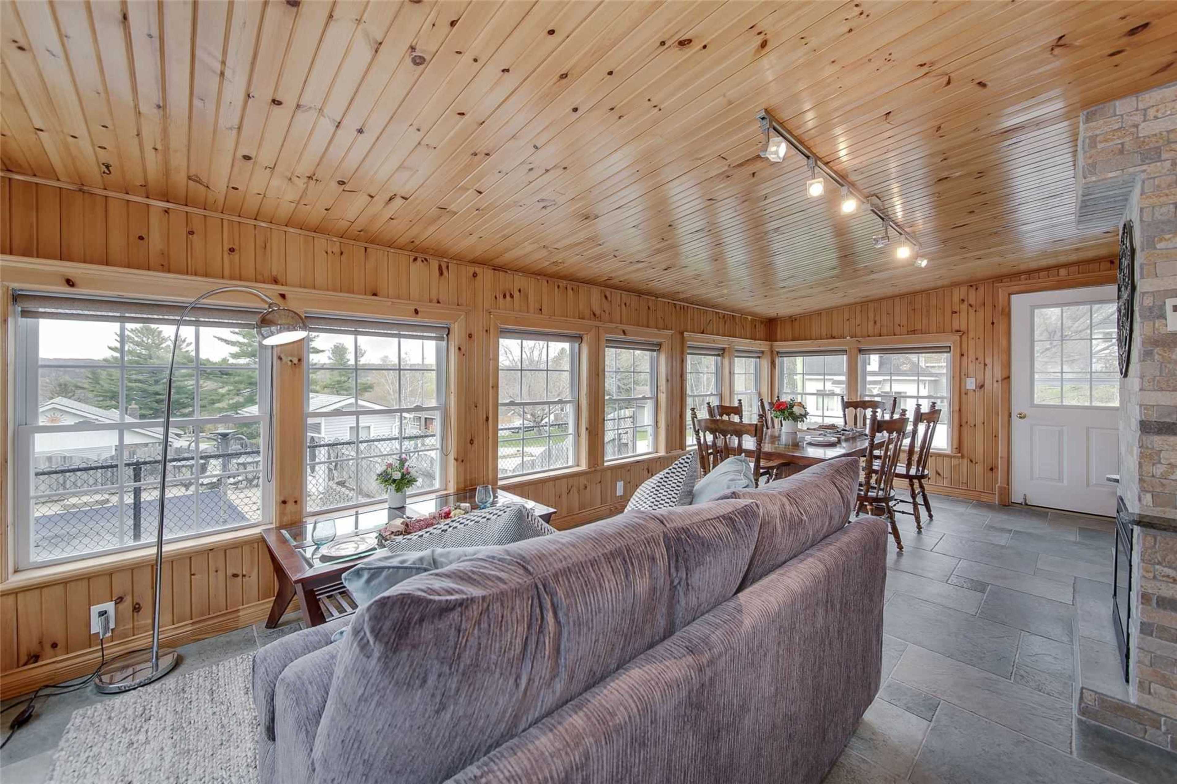 The image shows a sunlit room with wood-paneled walls, multiple windows, and a seating area featuring a gray sofa and wooden dining chairs.