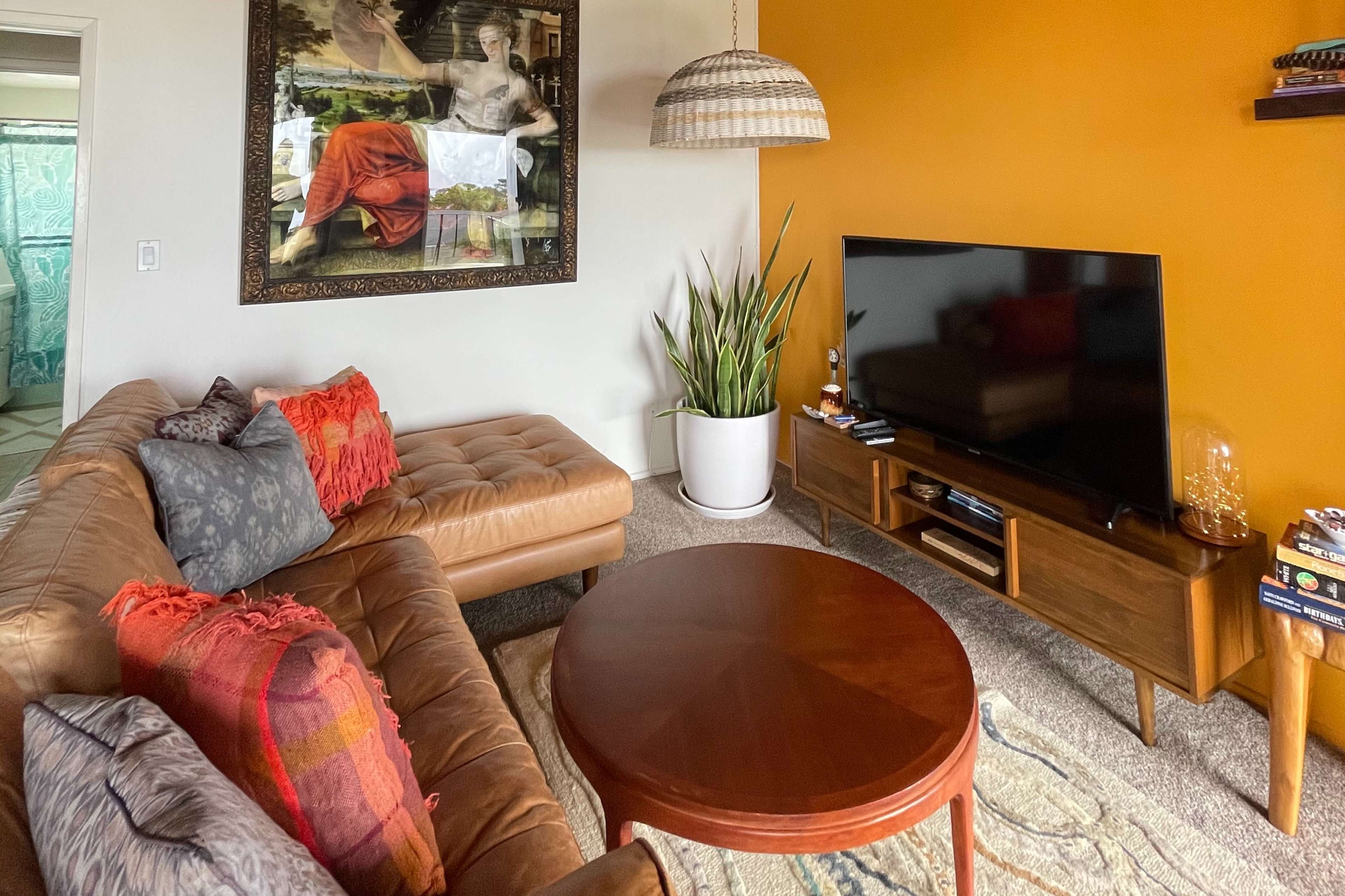 A modern living room features a brown leather sectional sofa, a round wooden coffee table, a large television mounted on a console, and a vibrant orange accent wall with a painting overhead.