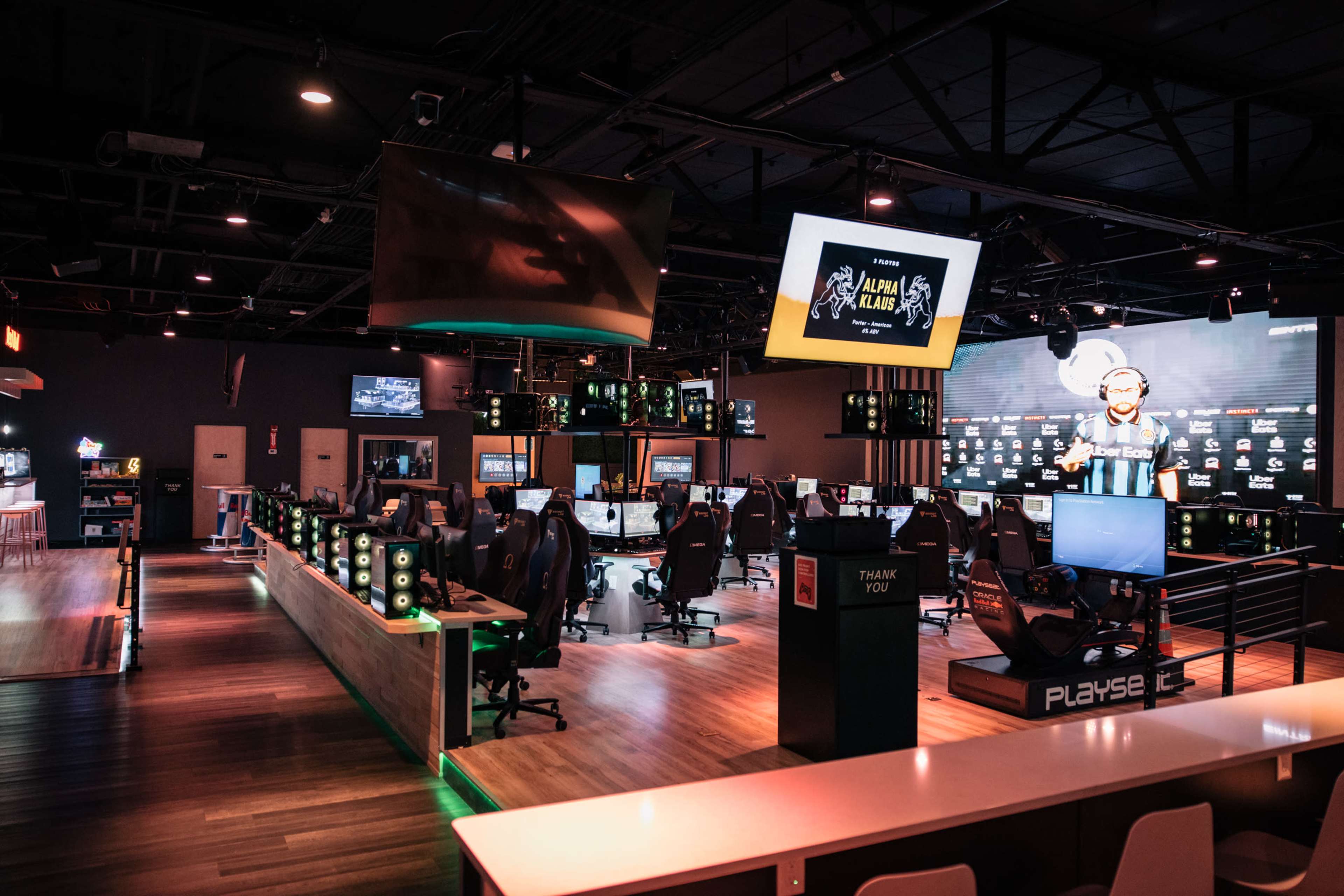 A modern esports gaming lounge features rows of high-end gaming computers, large screens displaying a tournament, and illuminated gaming accessories.