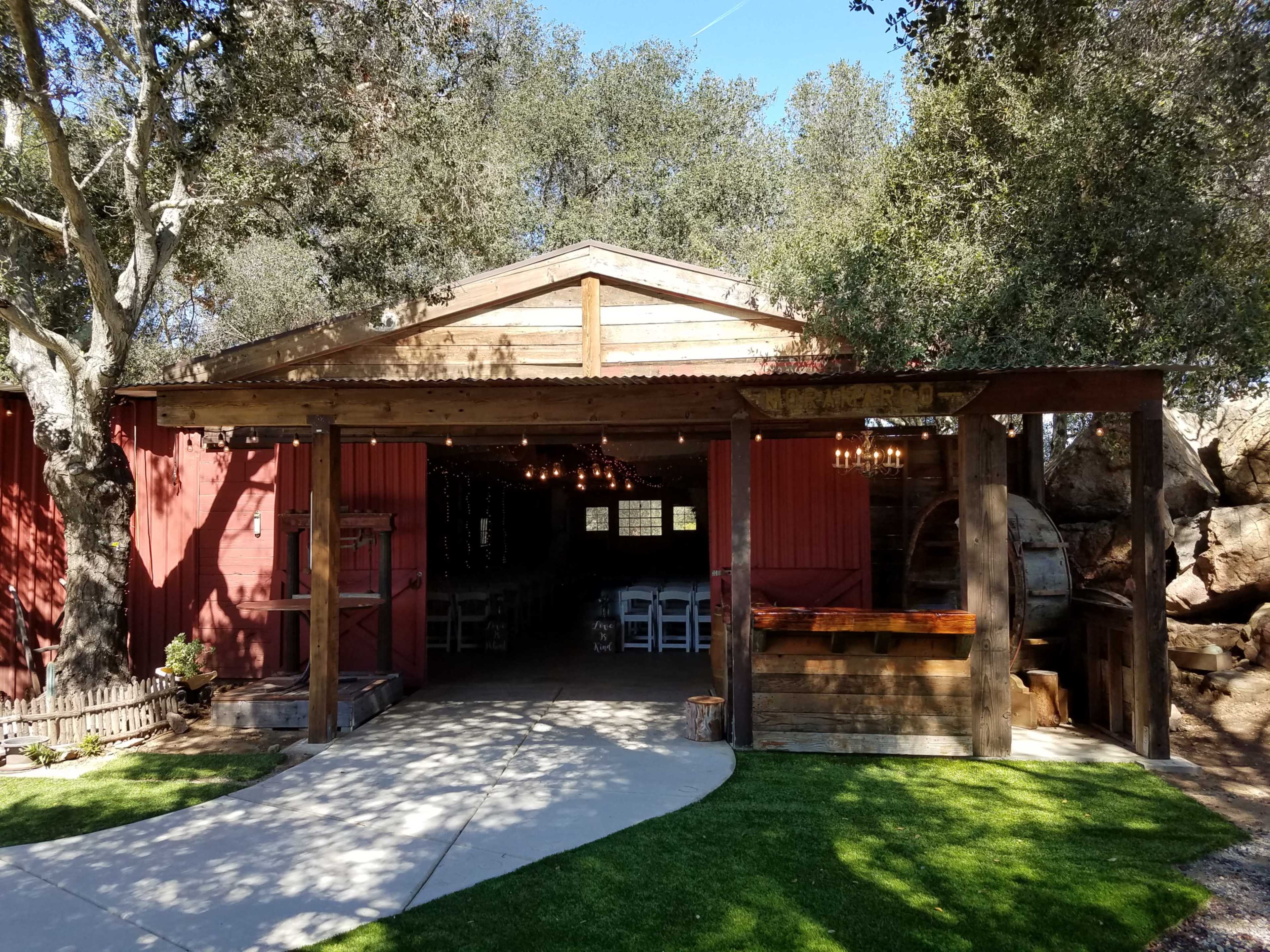 A rustic wooden structure with a corrugated metal roof features an open entrance, surrounded by trees and a manicured lawn.