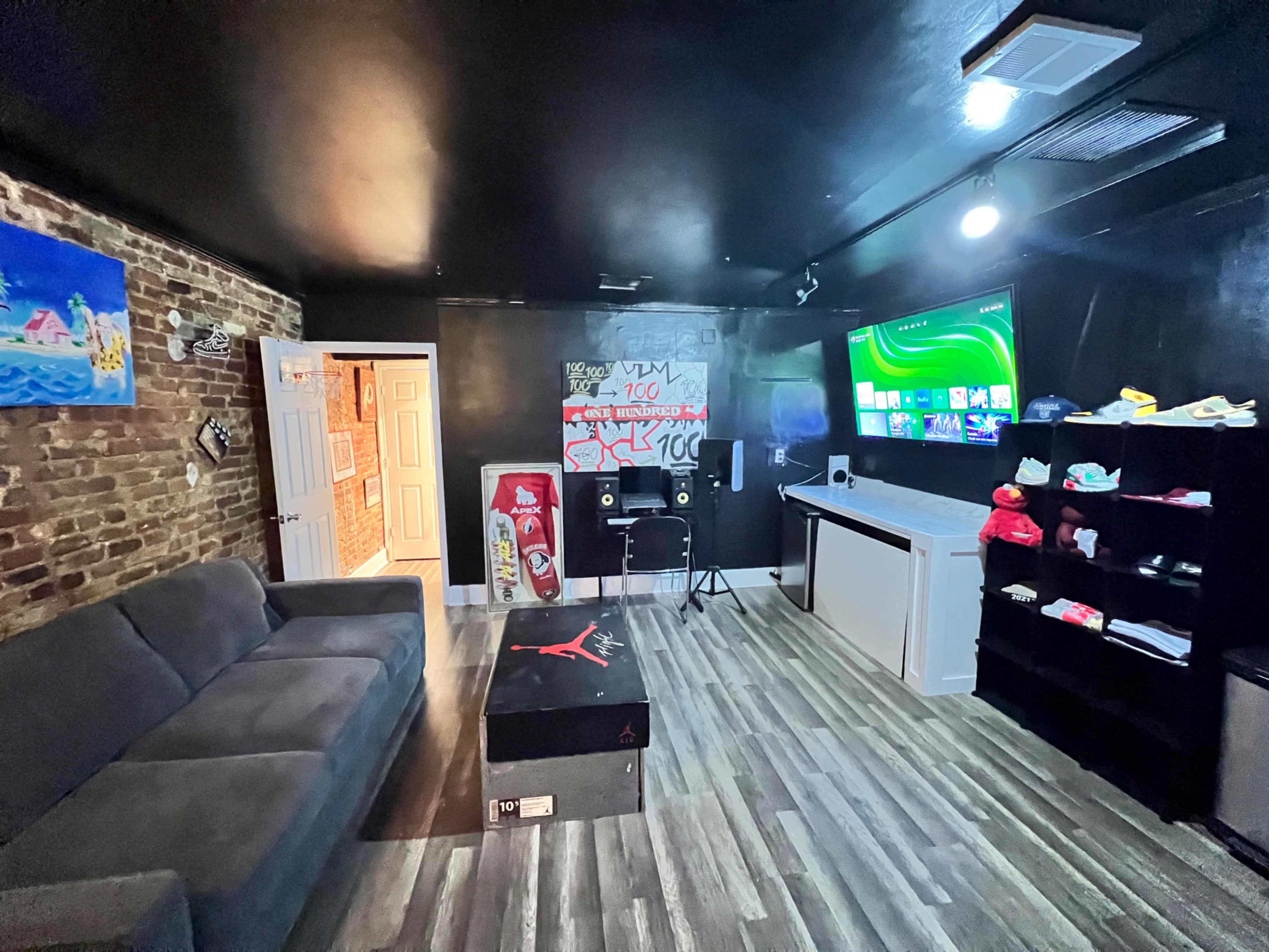 A dimly lit room with a black ceiling and walls, featuring a couch, a TV mounted on the wall, speakers, a storage shelf with clothing, and exposed brick on one side.