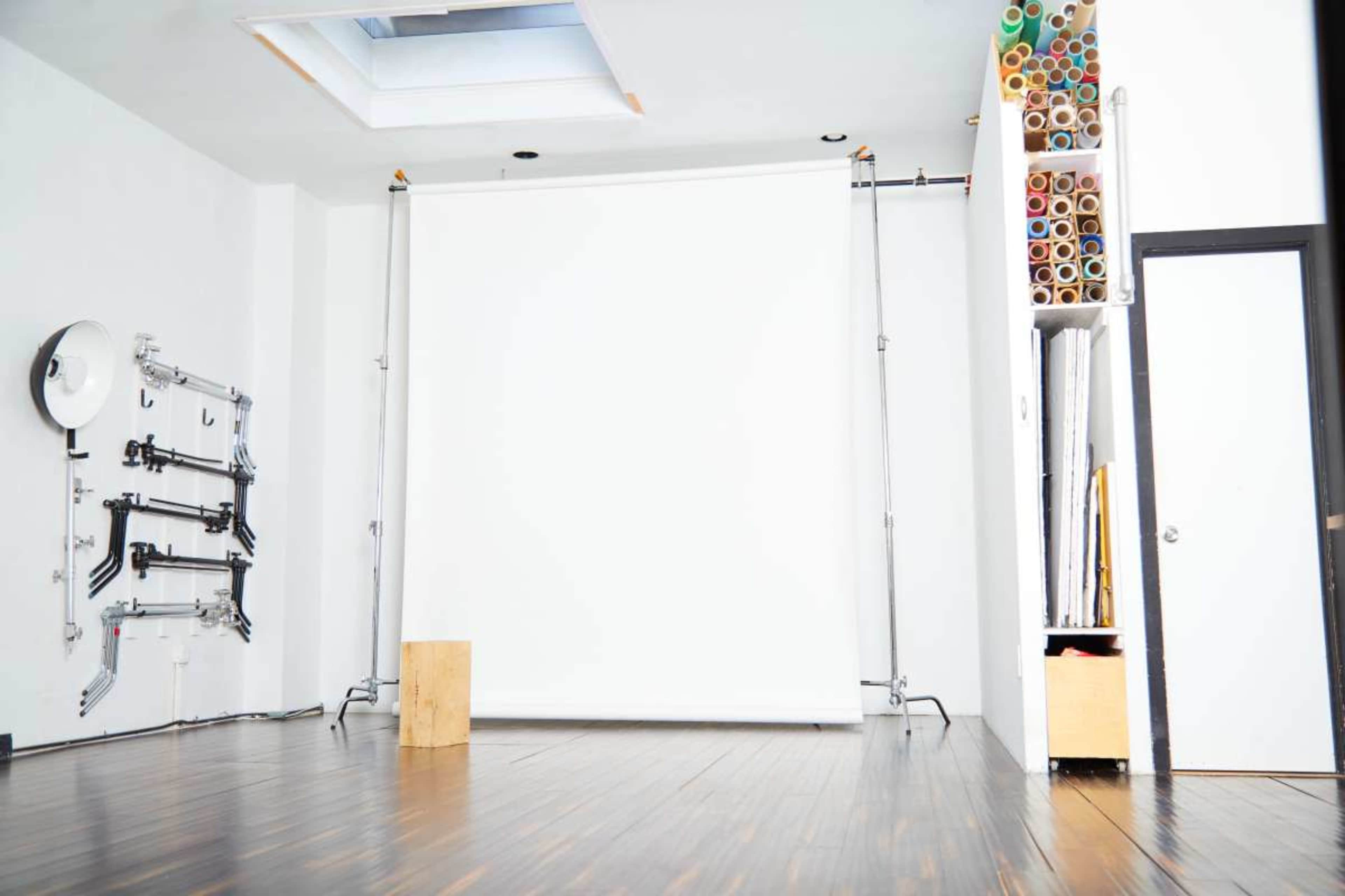 The image shows a bright, minimalistic studio space with a white backdrop, wooden flooring, and a storage area for rolled materials.