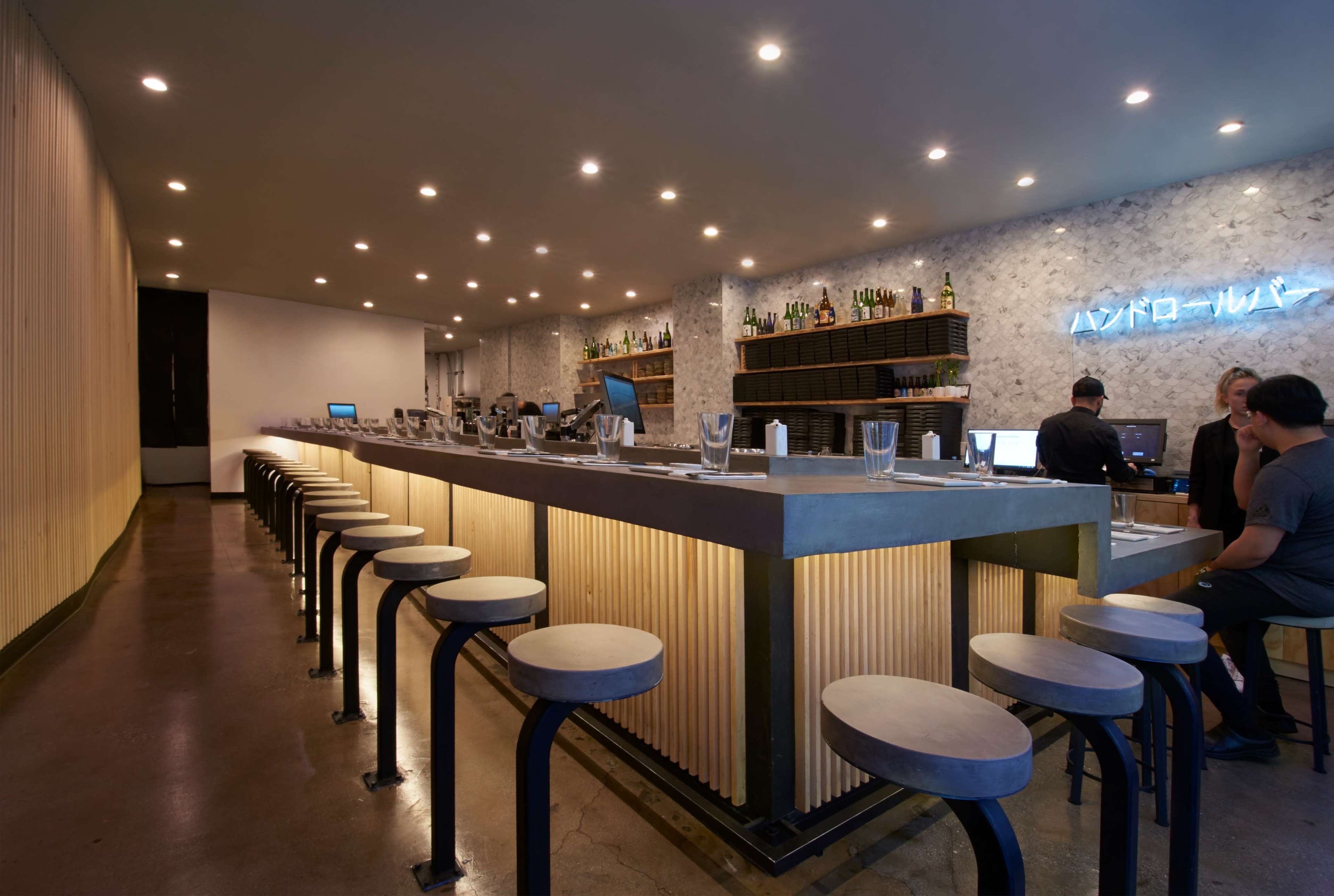 A modern bar interior with a long concrete counter, high stools, and warm lighting, complemented by a wall of bottles in the background.