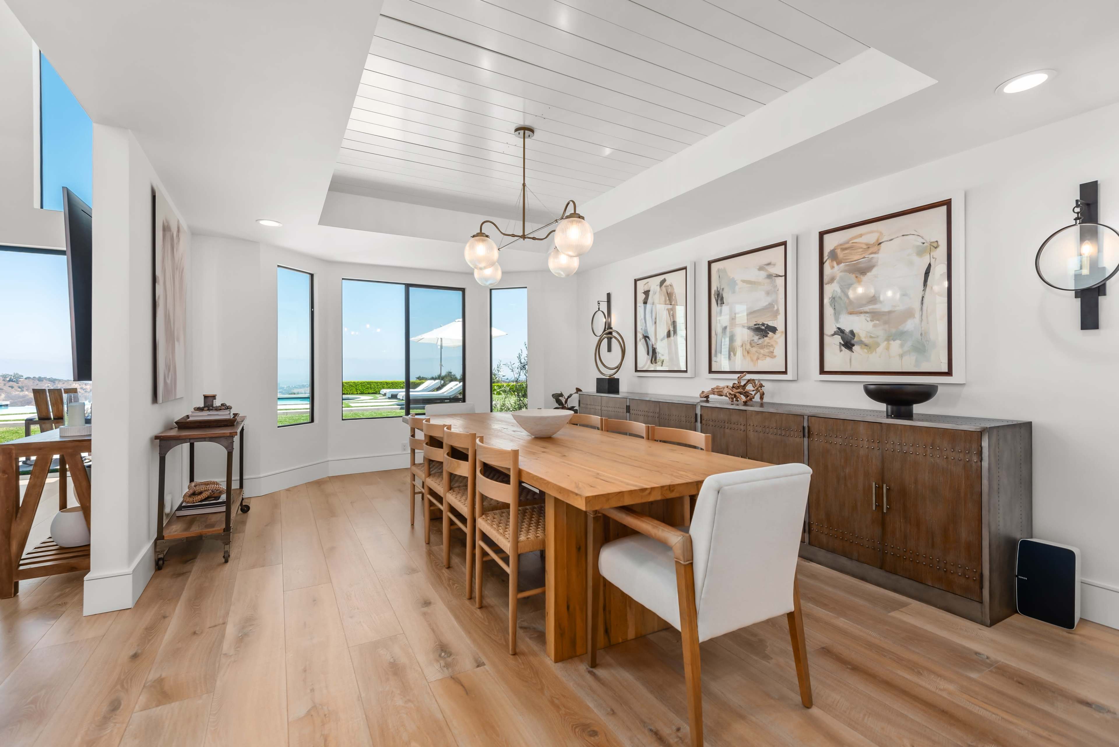 A modern dining room features a wooden table surrounded by chairs, with large windows showcasing a view of the outdoors and framed artwork on the walls.