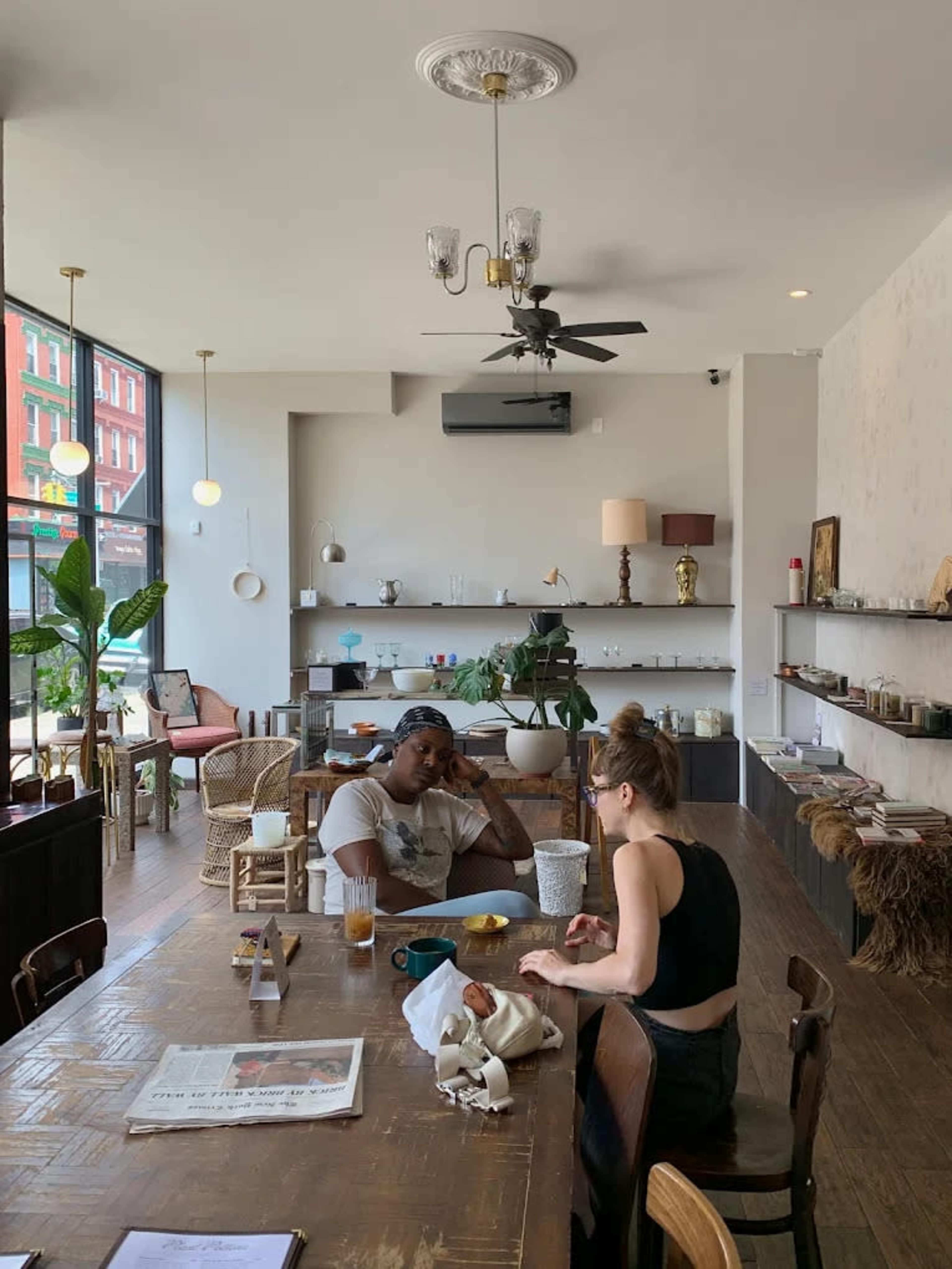 A man and a woman are seated at a large wooden table in a bright café, with various plants and decorative items arranged around them.