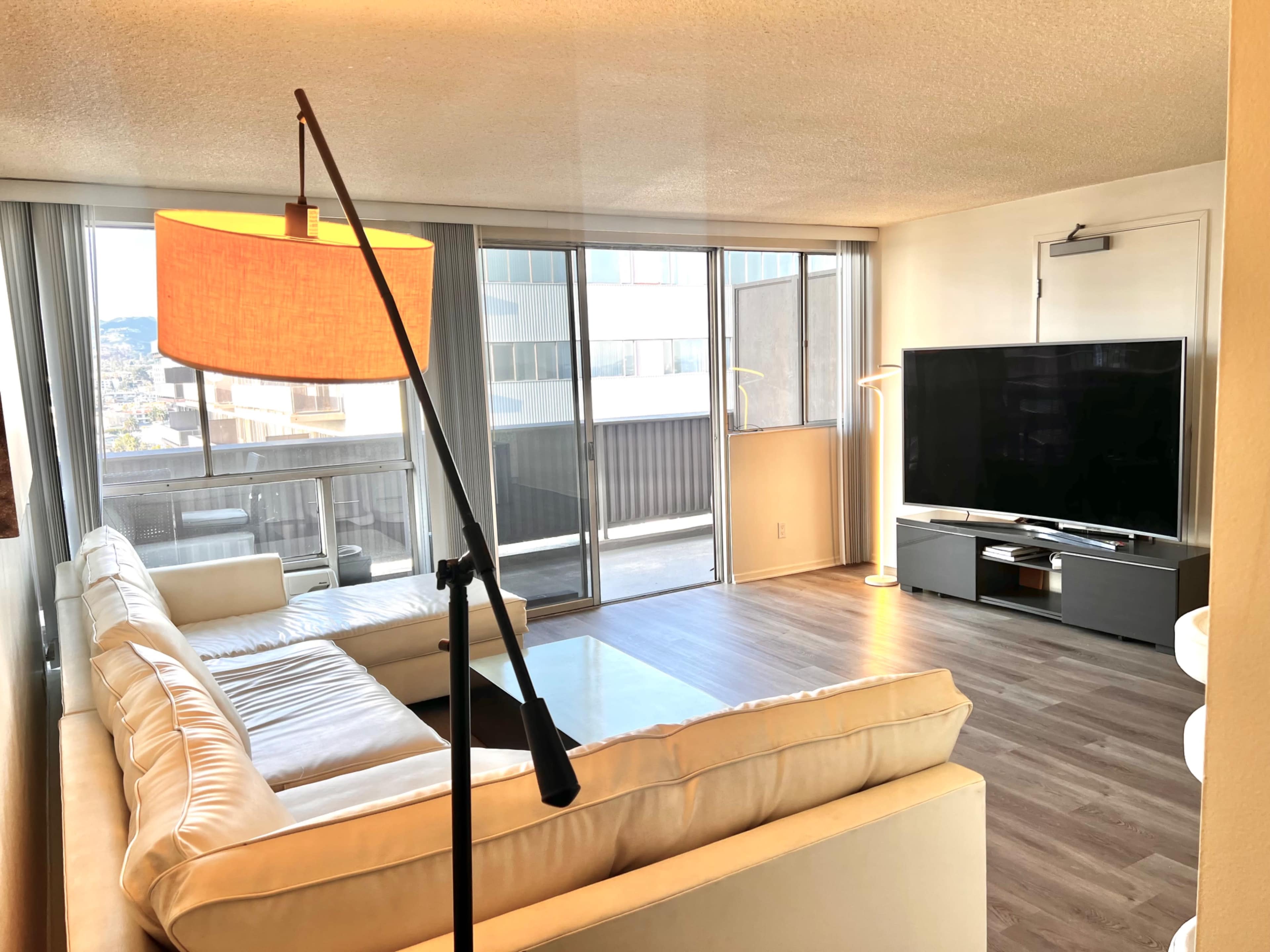 The image shows a modern living room with a large white sectional sofa, a flat-screen TV on a dark console, and large windows allowing natural light.