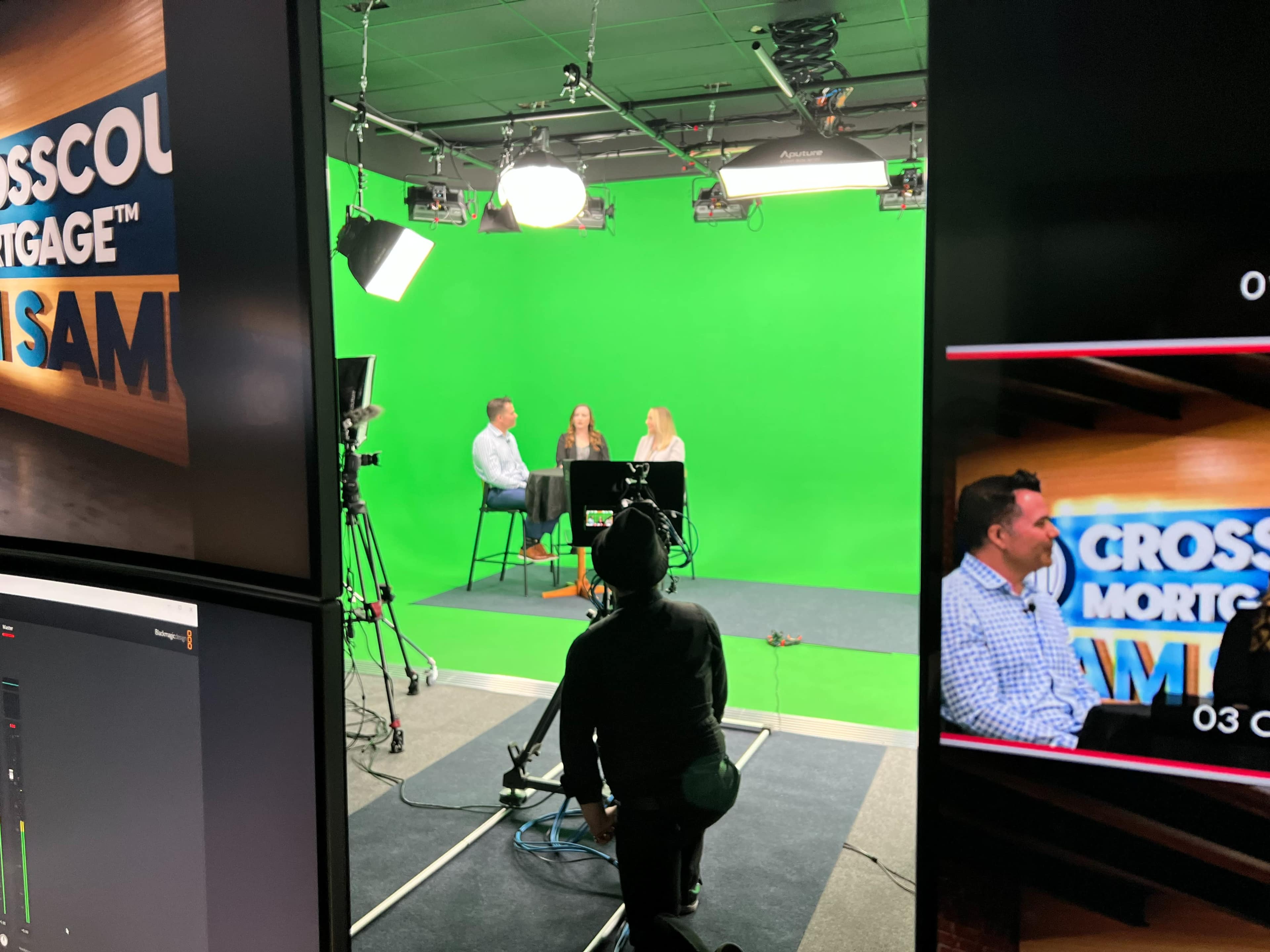A production set features two people seated on high chairs in front of a green screen, while a camera operator stands in the foreground.