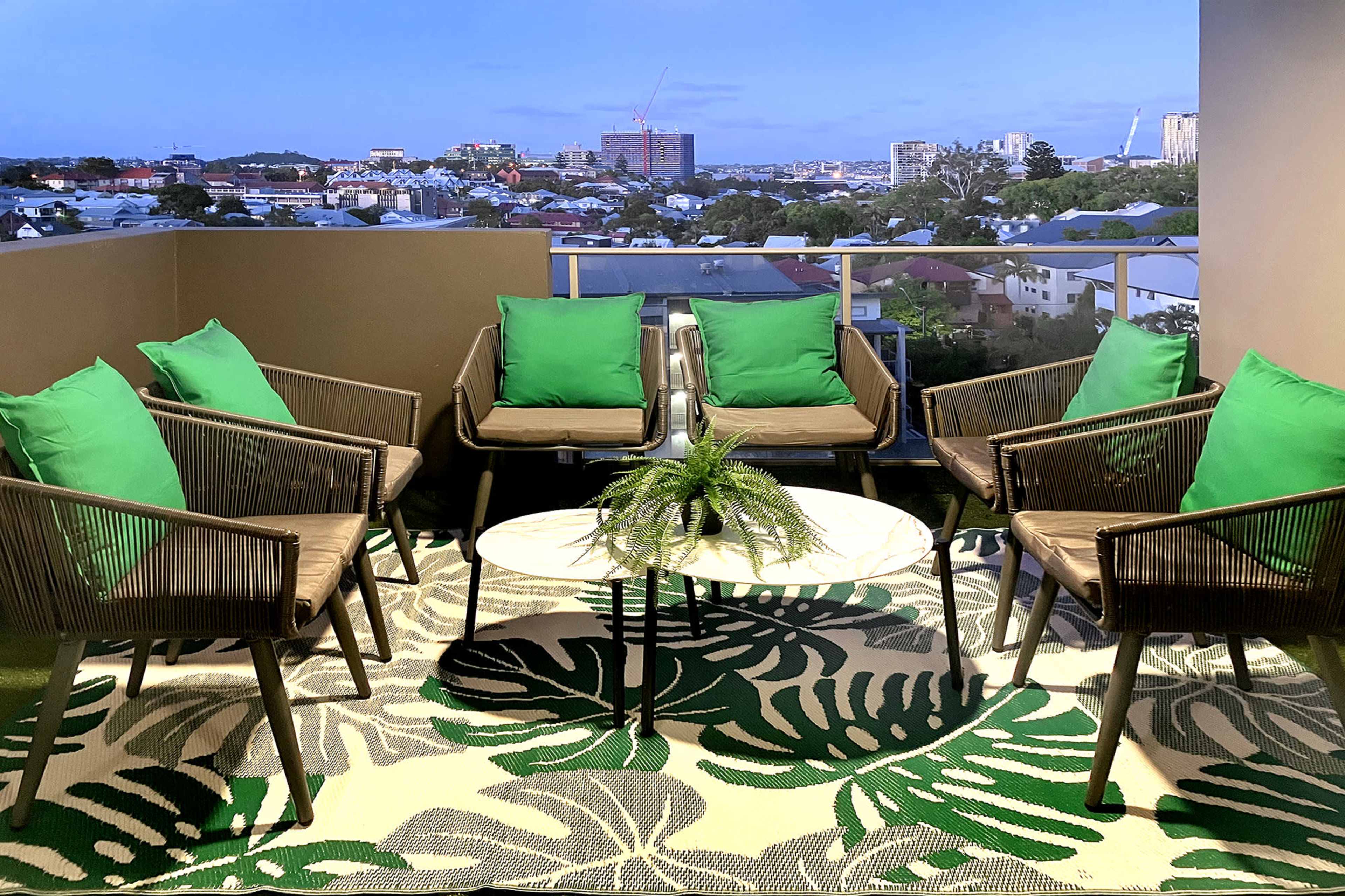 A balcony seating area features six chairs with green cushions arranged around a small table, overlooking a city skyline.