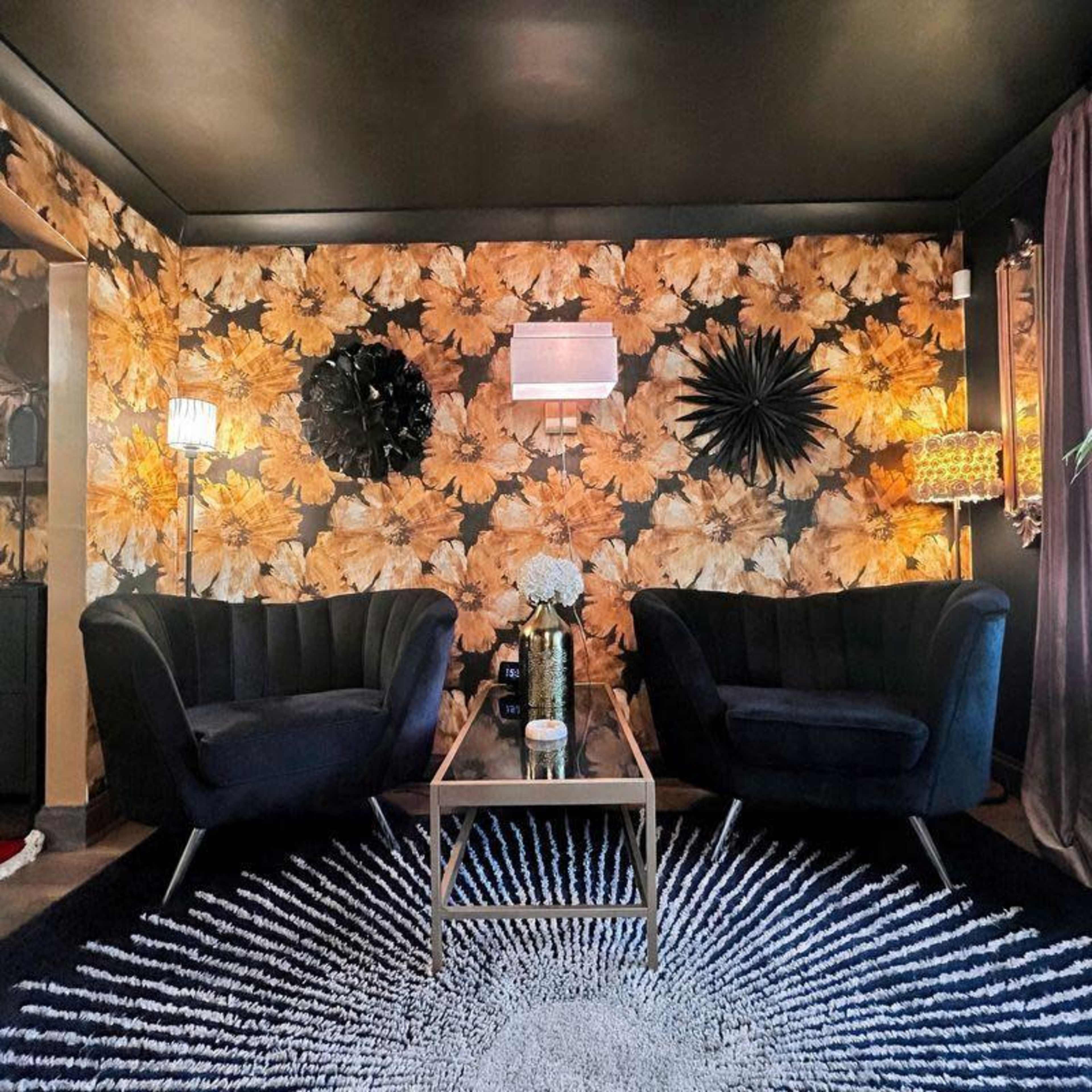 The scene features two modern velvet armchairs facing each other on a circular rug, with a coffee table in between, against a backdrop of floral wallpaper and various light fixtures.