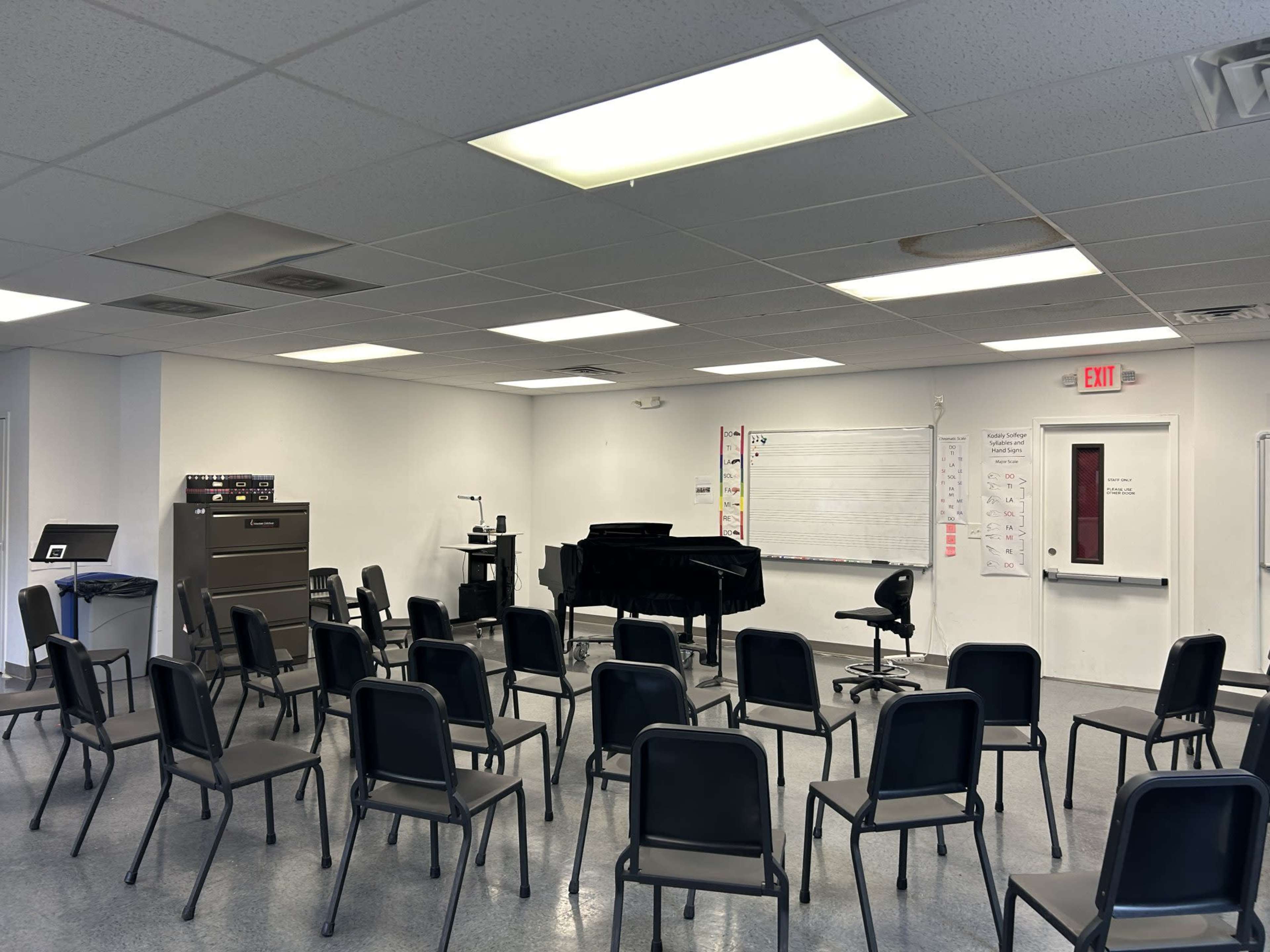 The image shows a music classroom with a grand piano, a whiteboard, and several rows of chairs arranged for a group setting.