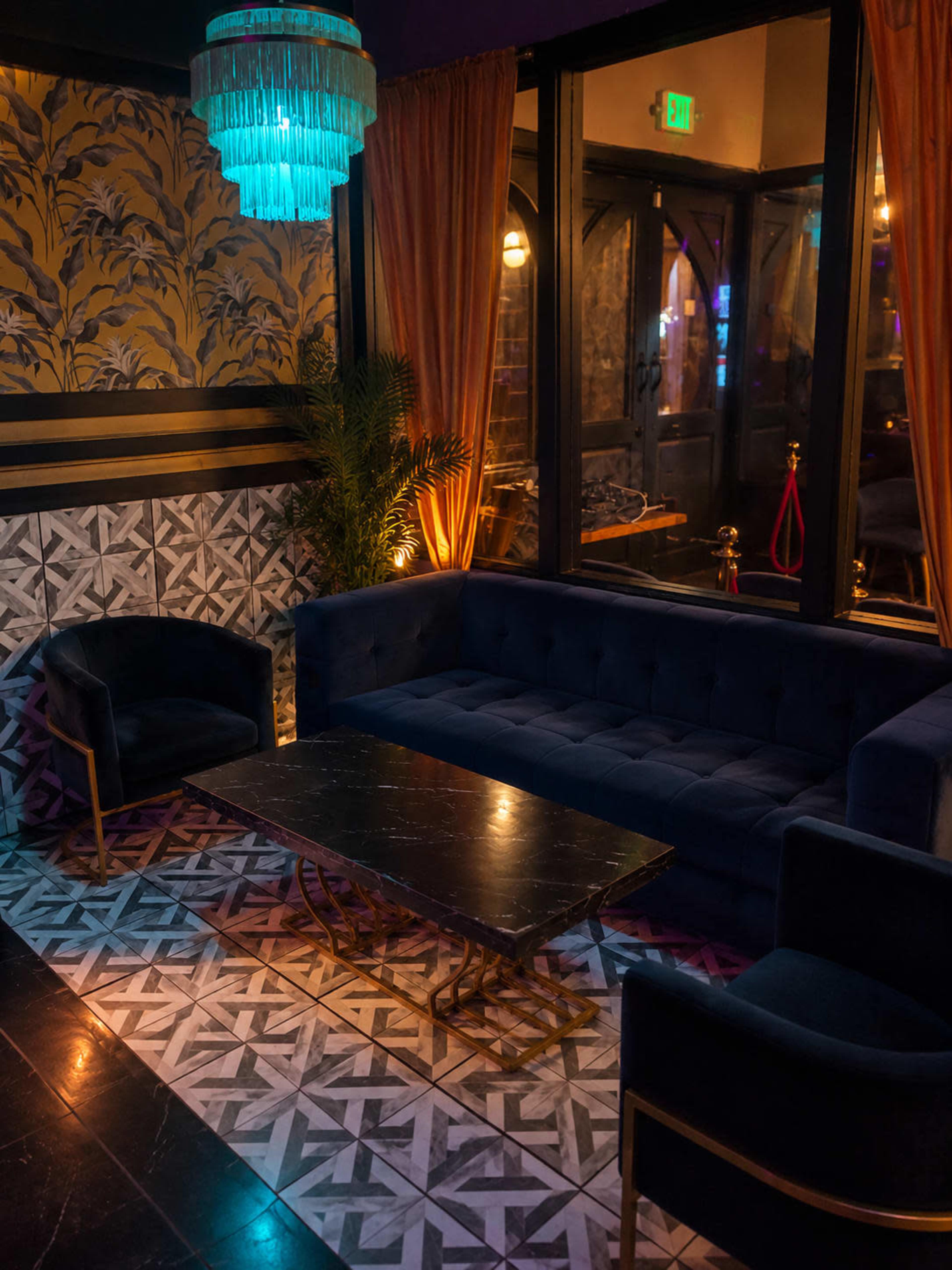 The image shows a cozy lounge area with a blue velvet sofa, two armchairs, and a marble-topped coffee table, illuminated by a turquoise chandelier.